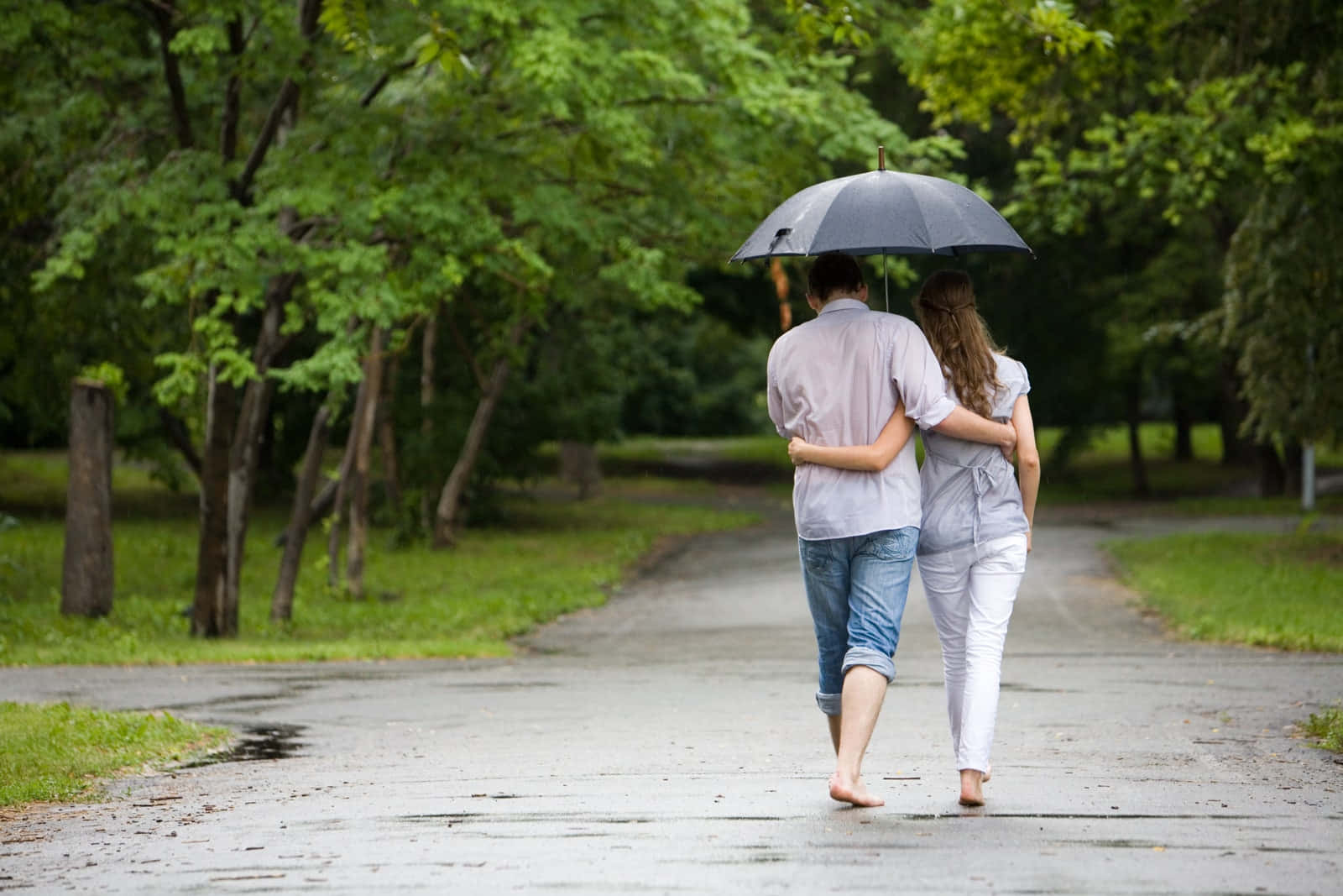 愛在雨中同行，堅守彼此 壁紙 — 一對情侶赤腳撐傘走在潮濕的公園小徑上，周圍是茂密的綠樹。