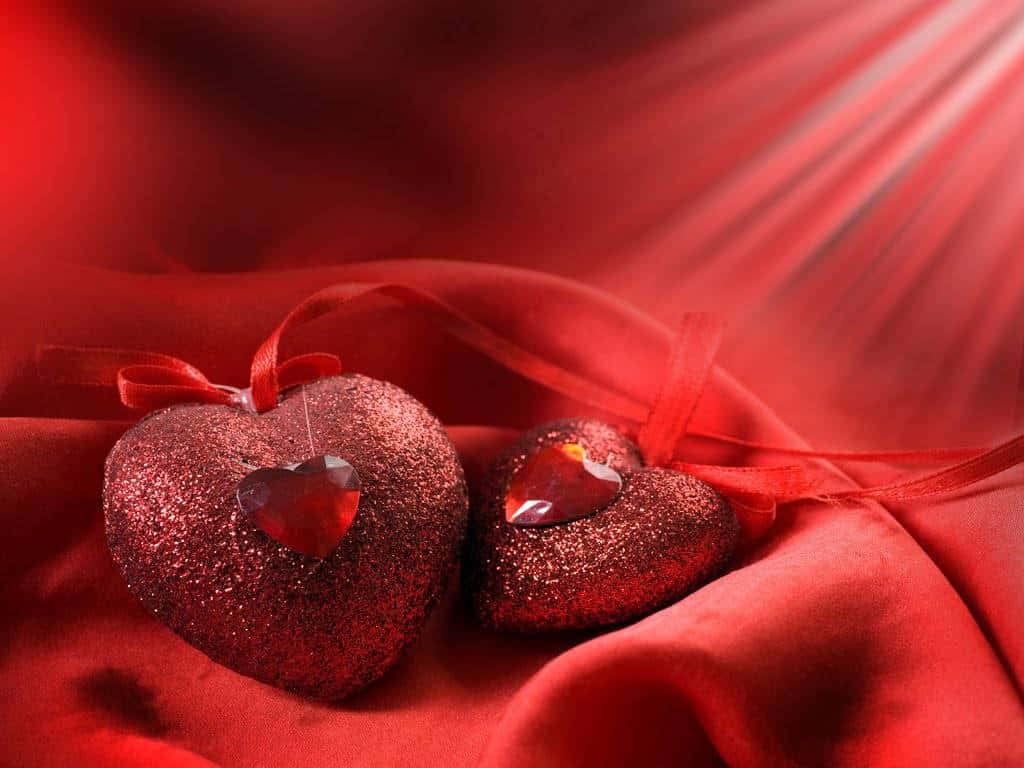 愛是如此甜美，一種難以言喻的美好體驗 壁紙 — 兩個閃閃發光的紅色心形裝飾品，帶有水晶中心，放置在溫暖光線沐浴下的流動紅色布料上。