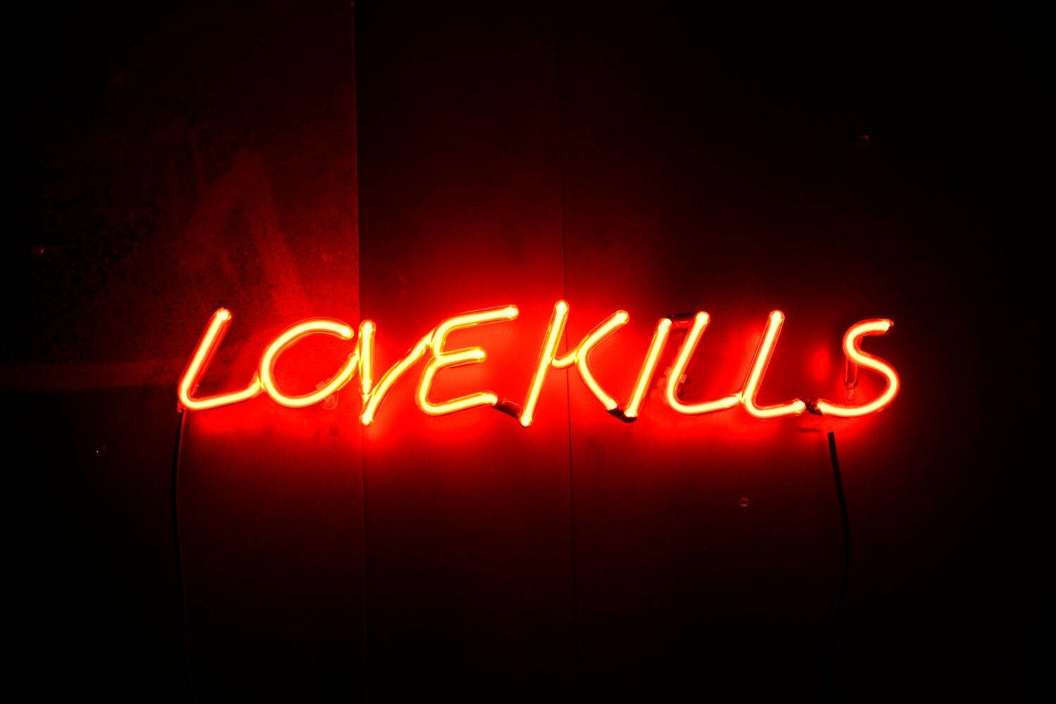 愛殺人語錄 壁紙 — 一個發光的紅色霓虹燈牌，拼寫著LOVEKILLS，在昏暗的空間中向深色紋理牆壁投射出溫暖的光芒。
