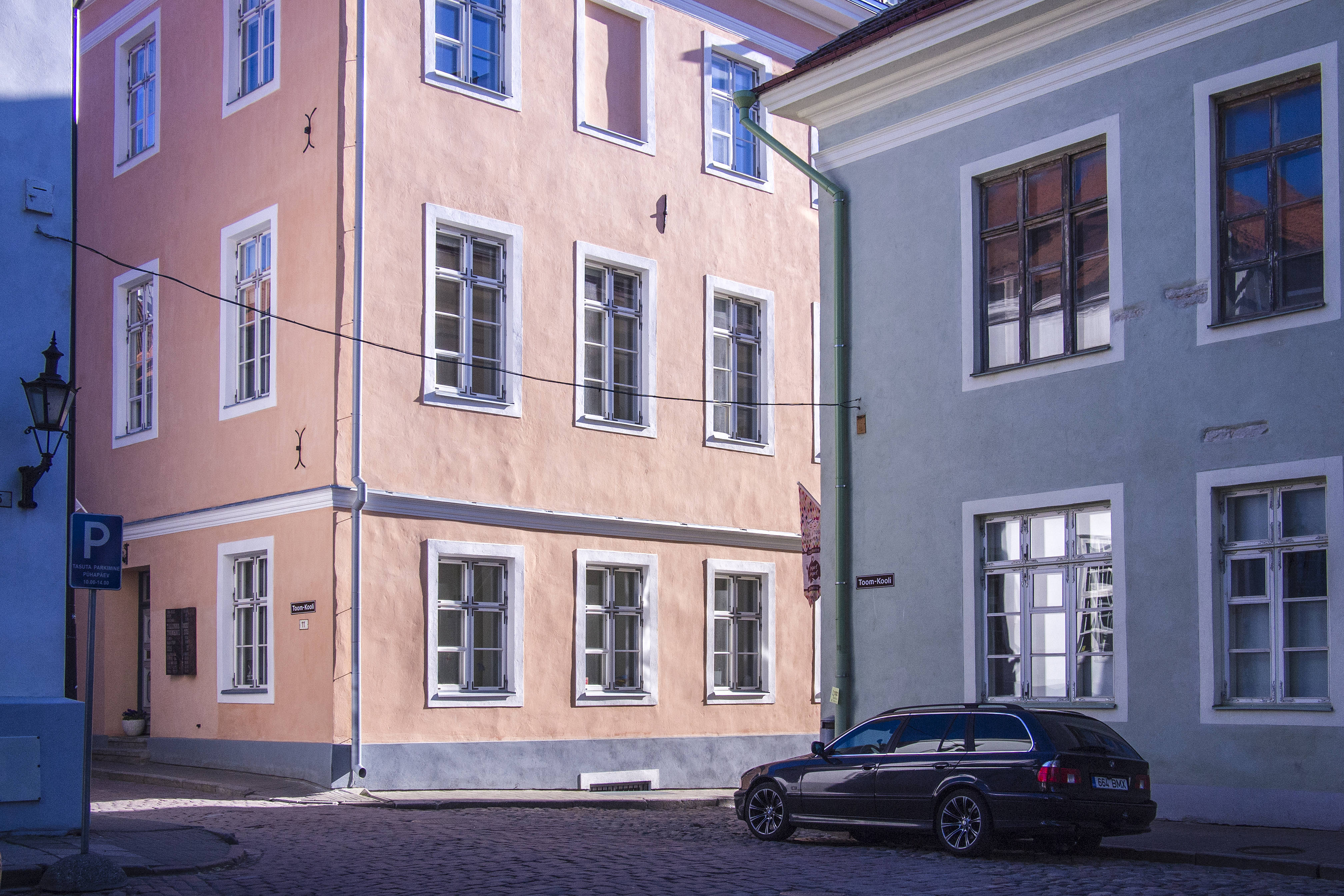愛沙尼亞施旺多夫城堡 壁紙 — 一個鵝卵石街道的角落，有粉彩色的建築物和一輛深色汽車在明亮的日光下停泊。