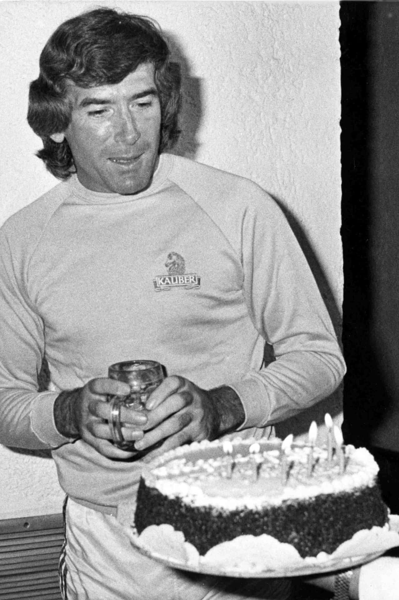 愛爾蘭足球傳奇 Pat Jennings 度過生日慶祝時刻 壁紙 — 一名穿著復古運動衫的男子在昏暗的房間裡，手持馬克杯，旁邊放著一個插著點燃蠟燭的生日蛋糕。