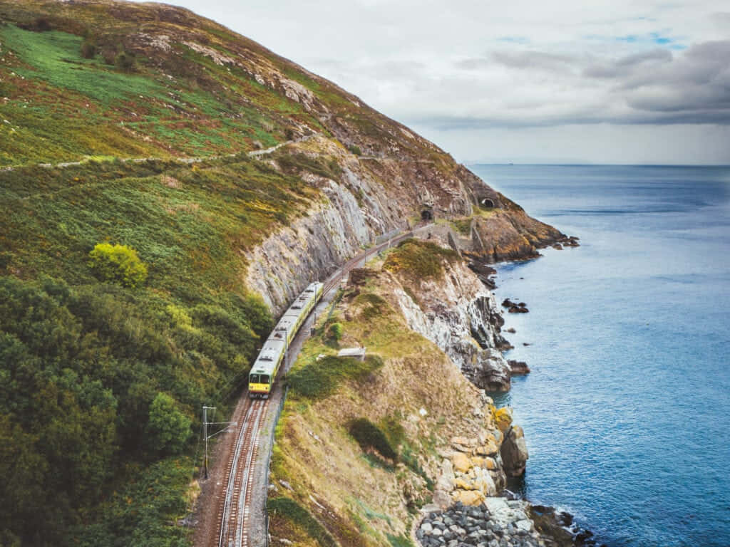 愛爾蘭鄉村的美麗風景 壁紙 — 一列黃色火車沿著崎嶇的海岸懸崖軌道蜿蜒而行，旁邊是深藍色的海洋，天空多雲。
