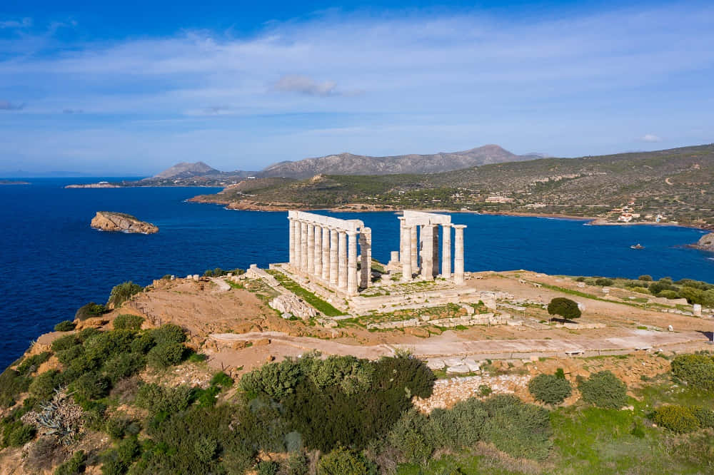 愛琴海與海神廟 壁紙 — 古代寺廟遺址，豎立的柱子棲息在懸崖上，俯瞰深藍色的大海和遠處的山脈，在明亮的天空下。
