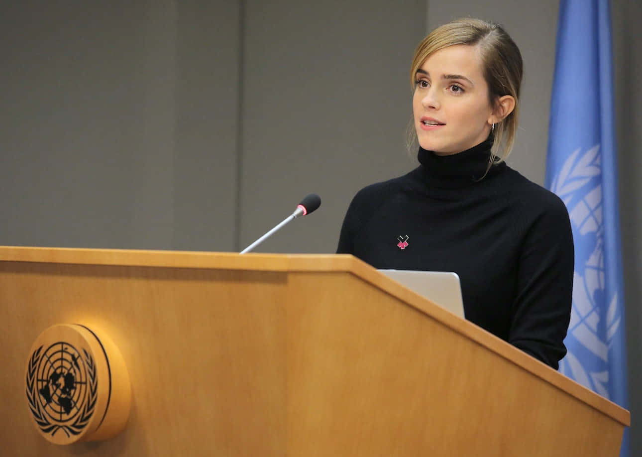 Emma Watson 桌布 壁紙 — 愛瑪屈臣穿著黑色高領毛衣，在藍色聯合國旗幟後方的木製聯合國講台上發言。