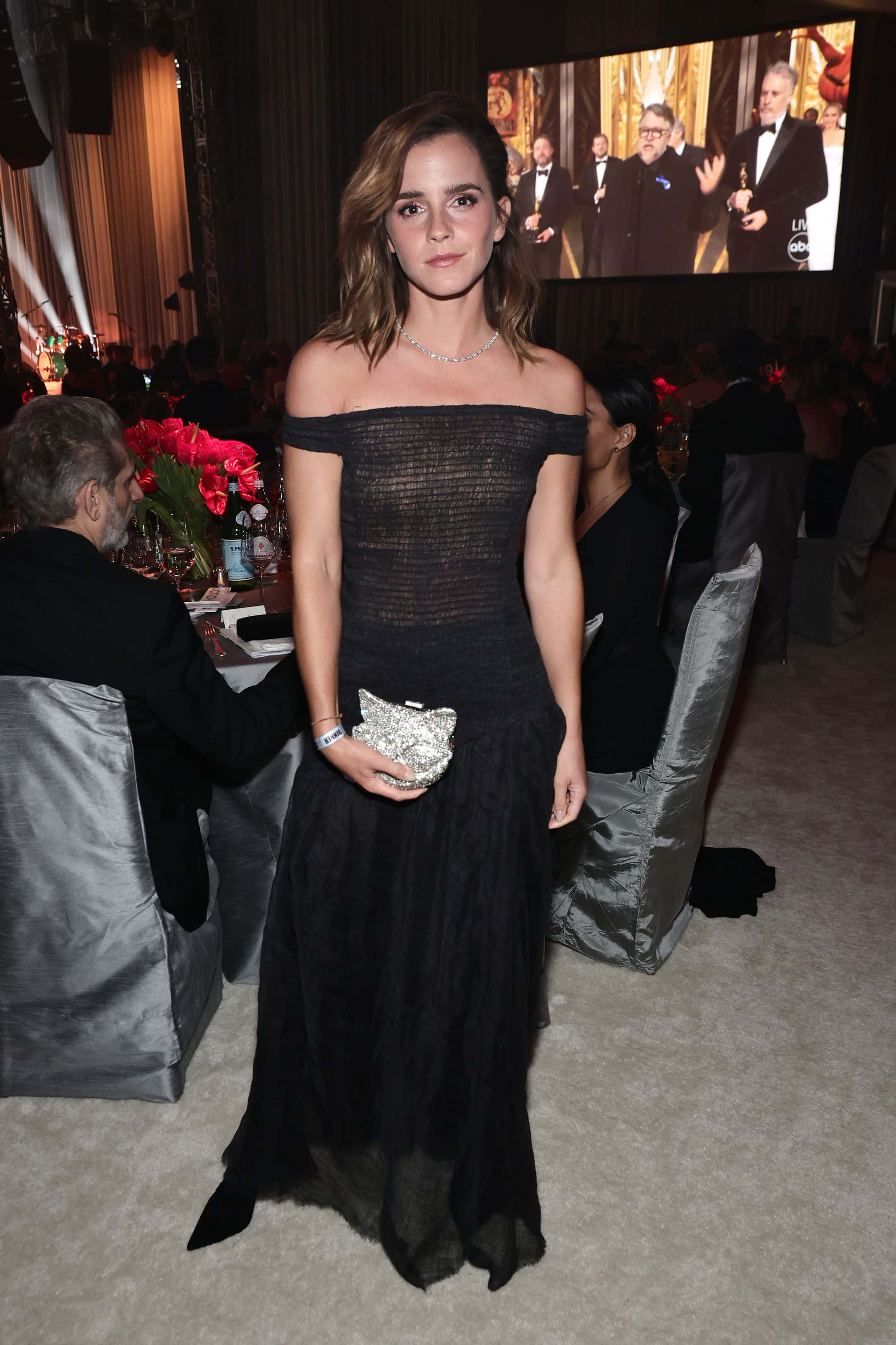 Emma Watson 桌布 壁紙 — 愛瑪屈臣身穿黑色露肩長裙，手持閃亮手拿包，在一個有賓客和大型屏幕的正式活動中優雅地站立。