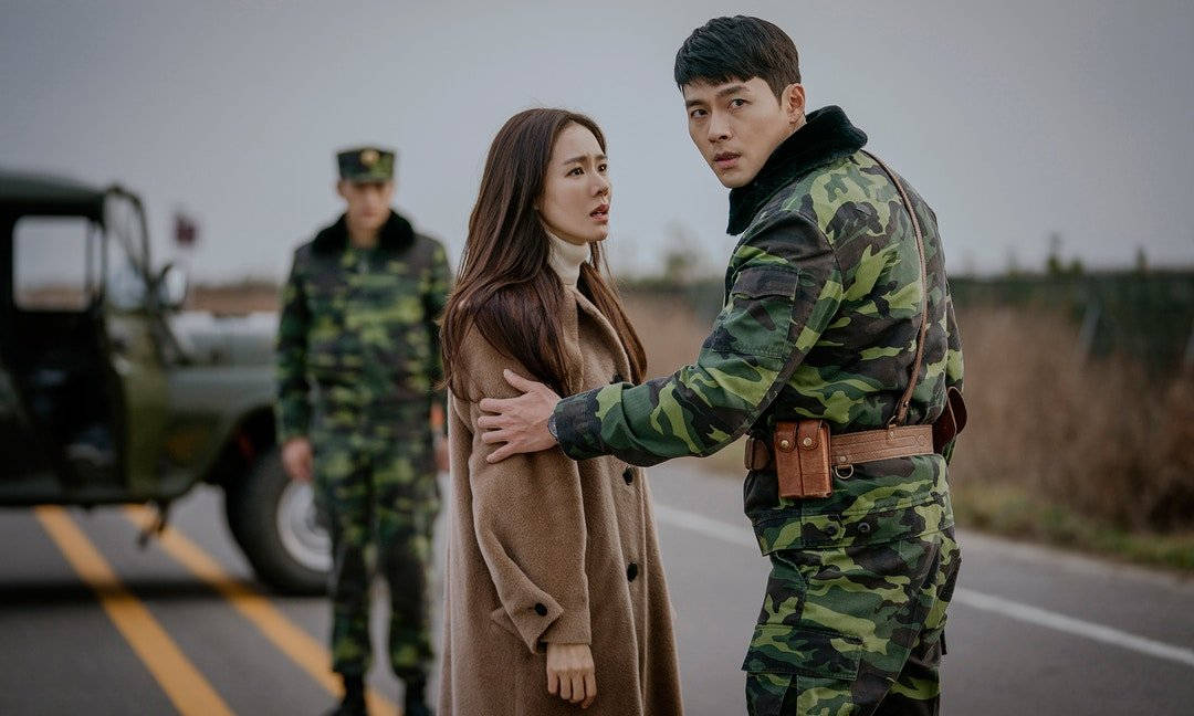 《愛的迫降》李政赫與世莉 壁紙 — 一名身穿迷彩服的士兵在路邊輕輕擁抱著一位身穿棕色外套的女子，另一名士兵則在附近站崗。
