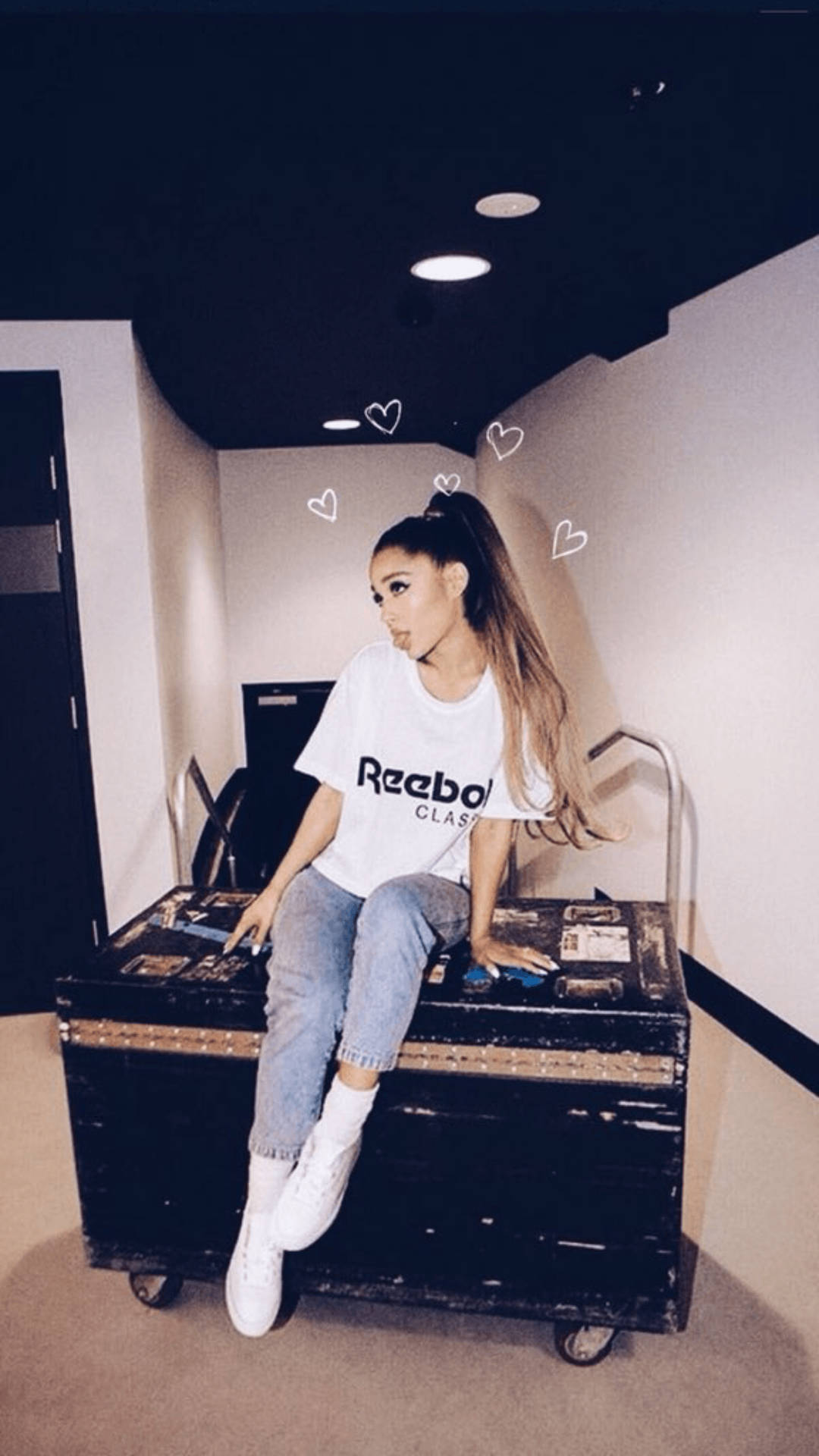 Ariana Grande 坐在黑色行李箱上，身穿白色 Reebok T 恤和牛仔褲，頭頂上方有心形塗鴉。