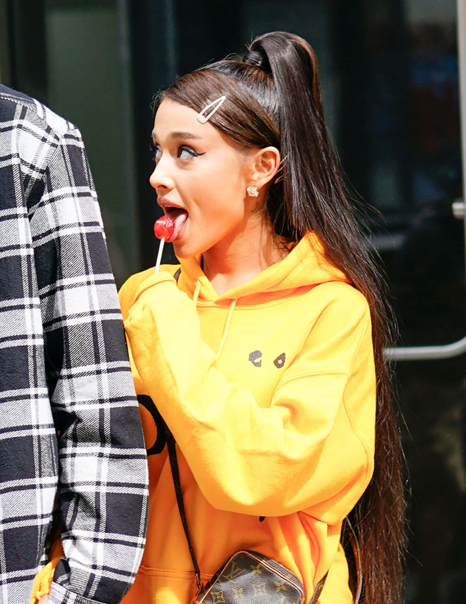 Ariana Grande 身穿黃色連帽衫，在戶外頑皮地舔著紅色棒棒糖。