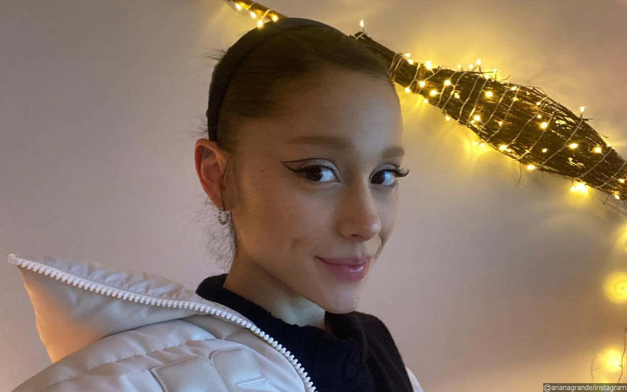 Ariana Grande 穿著白色外套，身後有仙女燈微笑。