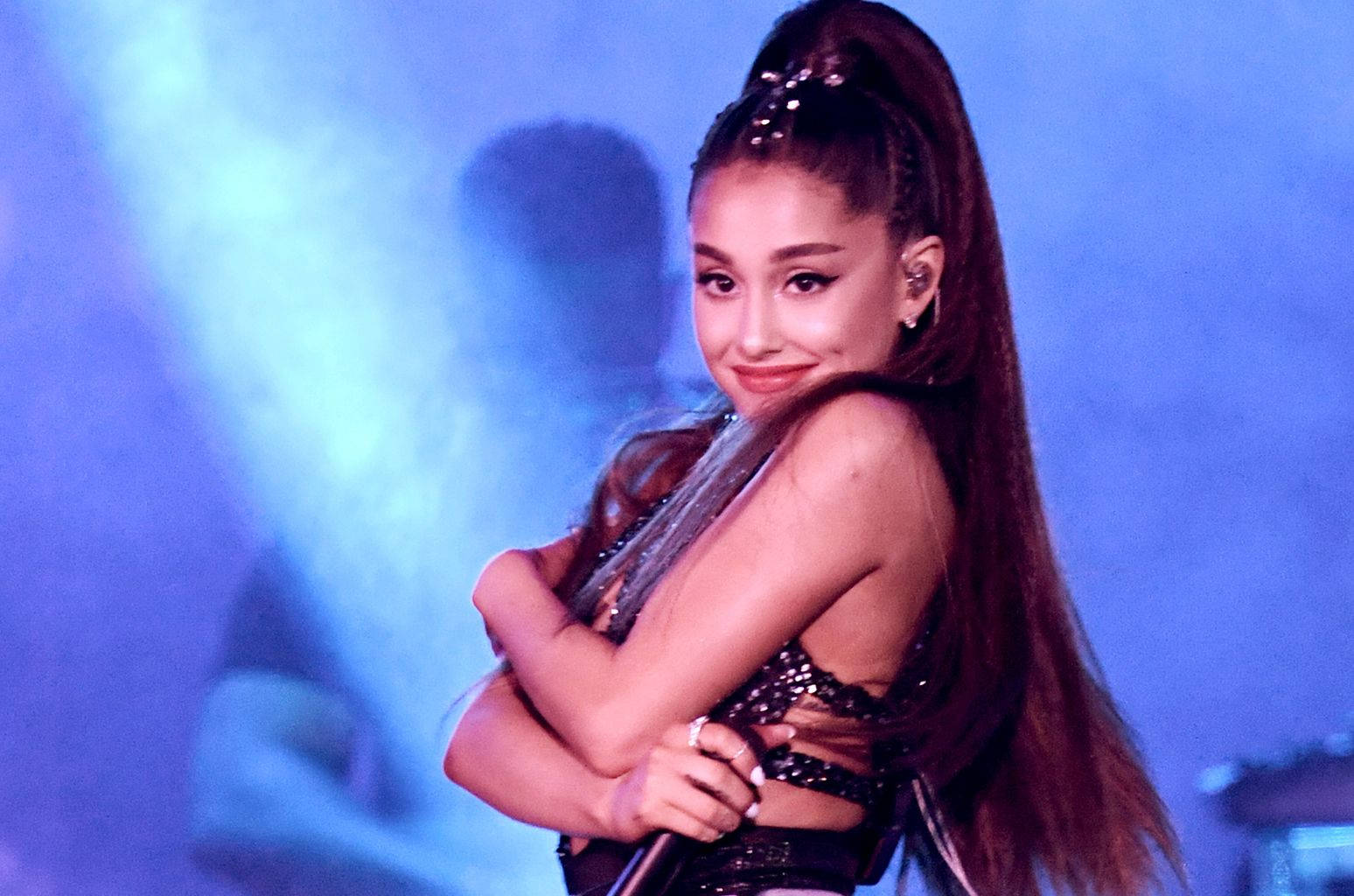 Ariana Grande 在藍色燈光下，身穿閃亮黑色服裝，在舞台上微笑。