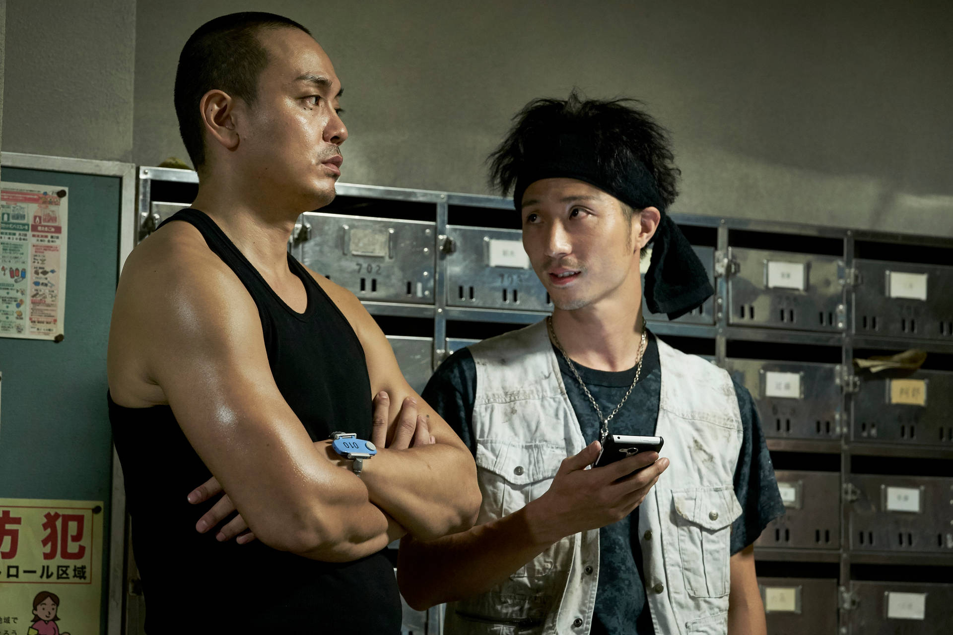 兩個男人在更衣室裡，一個雙手交叉，另一個拿著手機看起來很驚訝。
