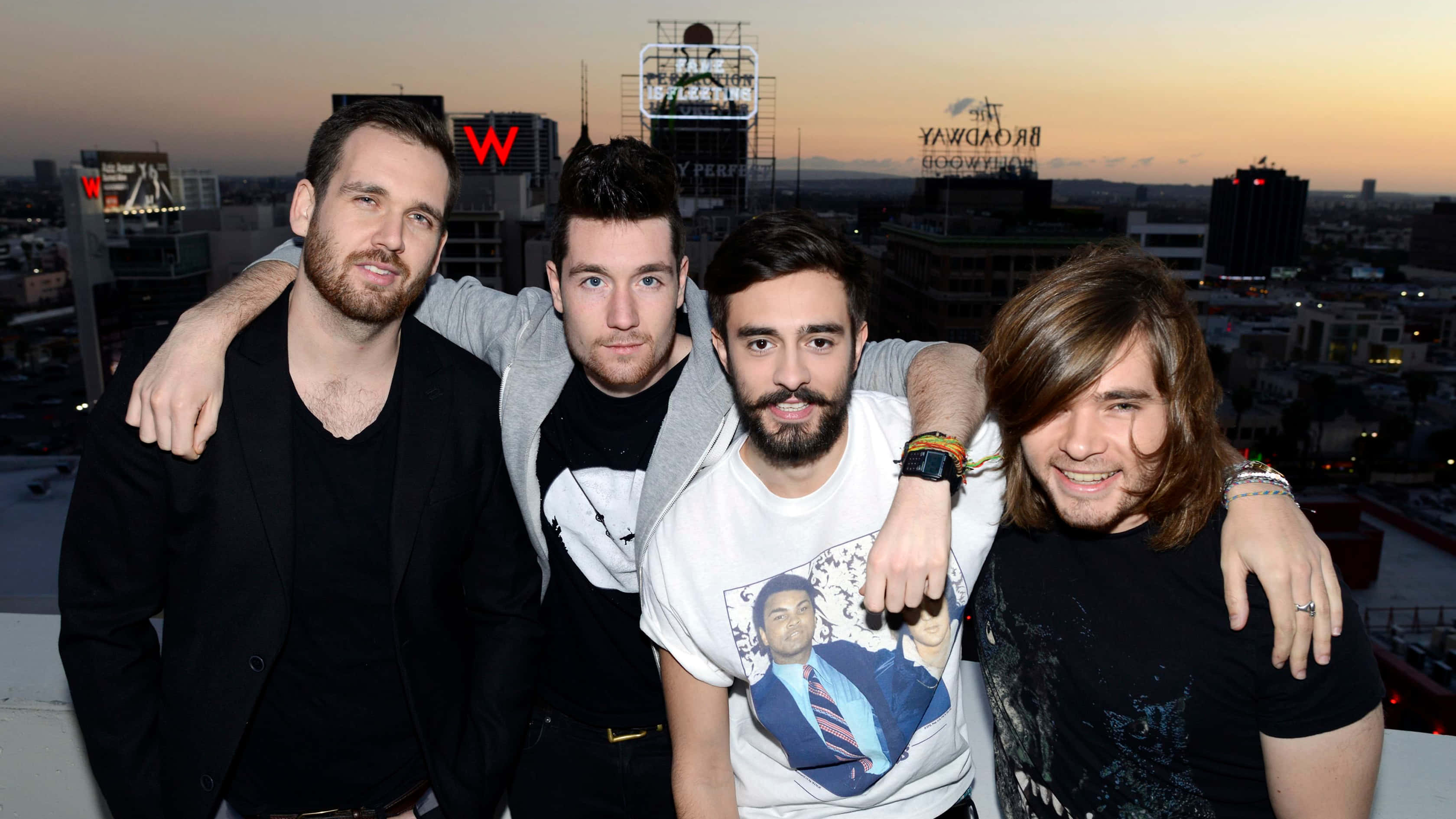 感受 Bastille 最新作品的澎湃能量 壁紙 — 四個男人在日落時份於天台一起擺姿勢，背後是城市天際線和發光的招牌。