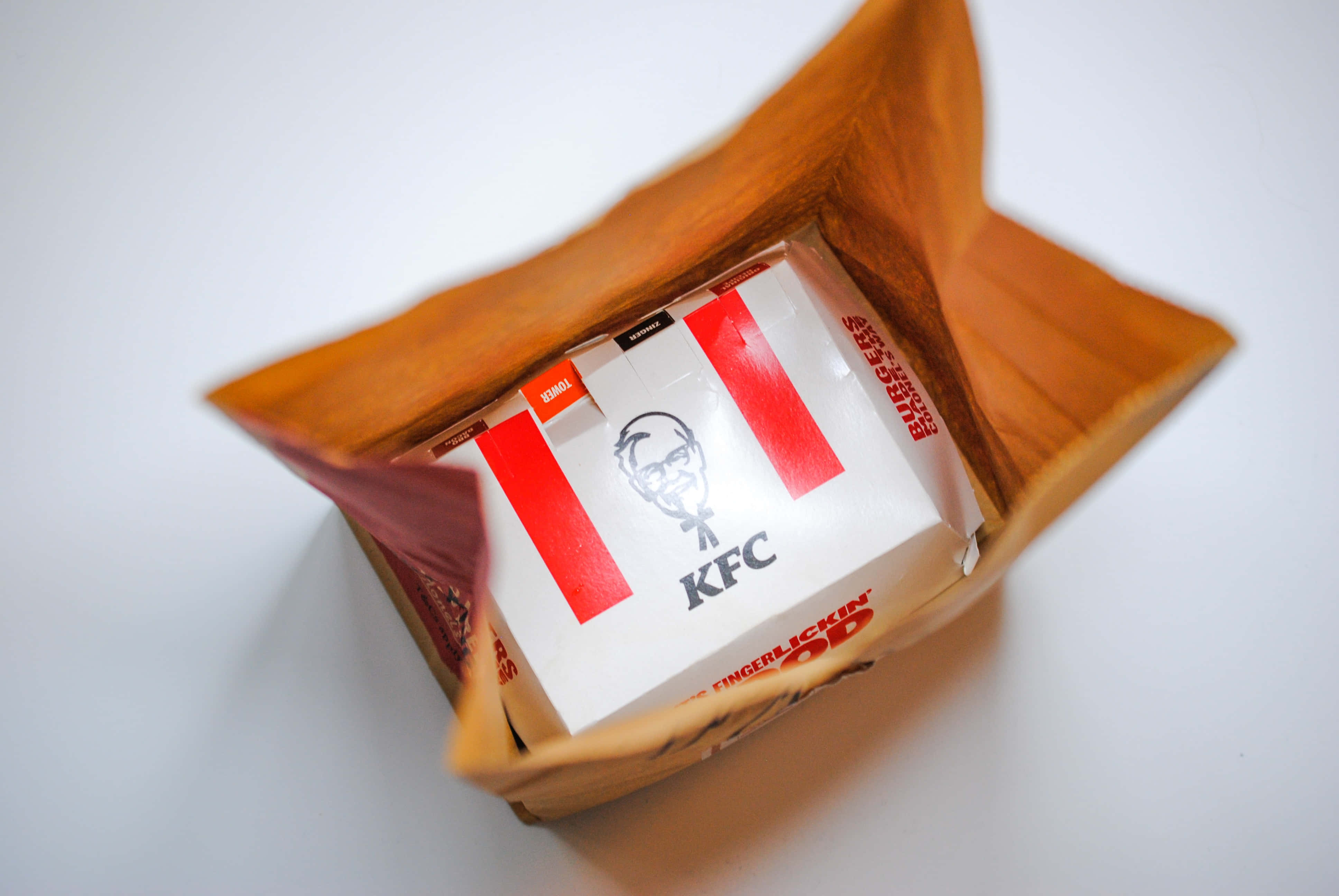 感受 KFC 真正的美味 壁紙 — 一個白色KFC盒，上面有紅色條紋，放在一個棕色紙袋內，置於純白色背景上的俯視圖。