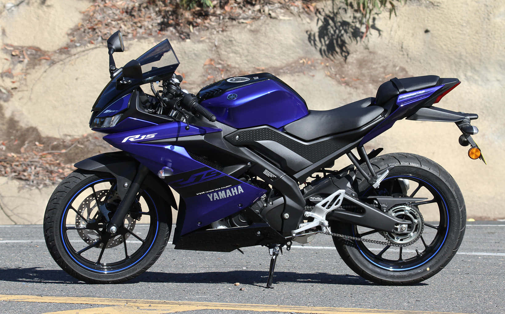 感受 R15 的強勁動力 壁紙 — 一架流線型藍色Yamaha R15電單車停泊在瀝青路上，背後有陽光照射的岩石牆。