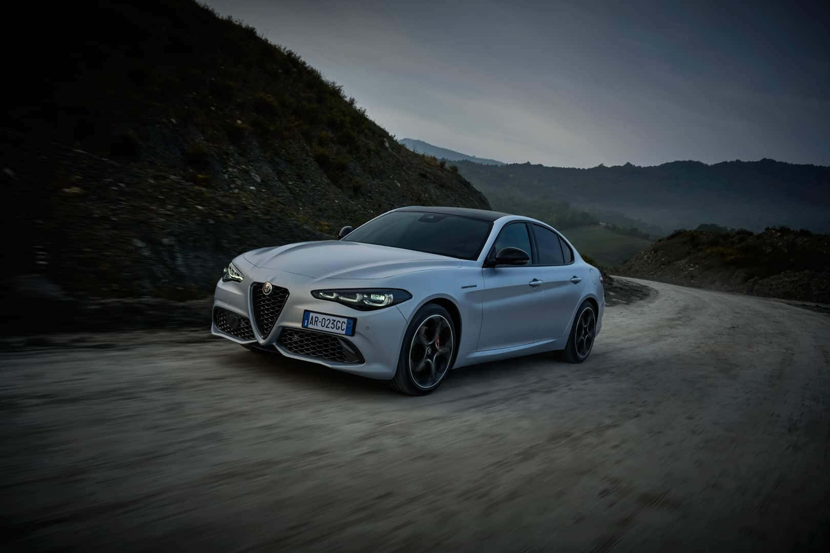 感受Alfa Romeo的澎湃動力 壁紙 — 一輛時尚的銀色 Alfa Romeo 轎車在蜿蜒的山路上疾馳，天空籠罩著暮色氛圍。