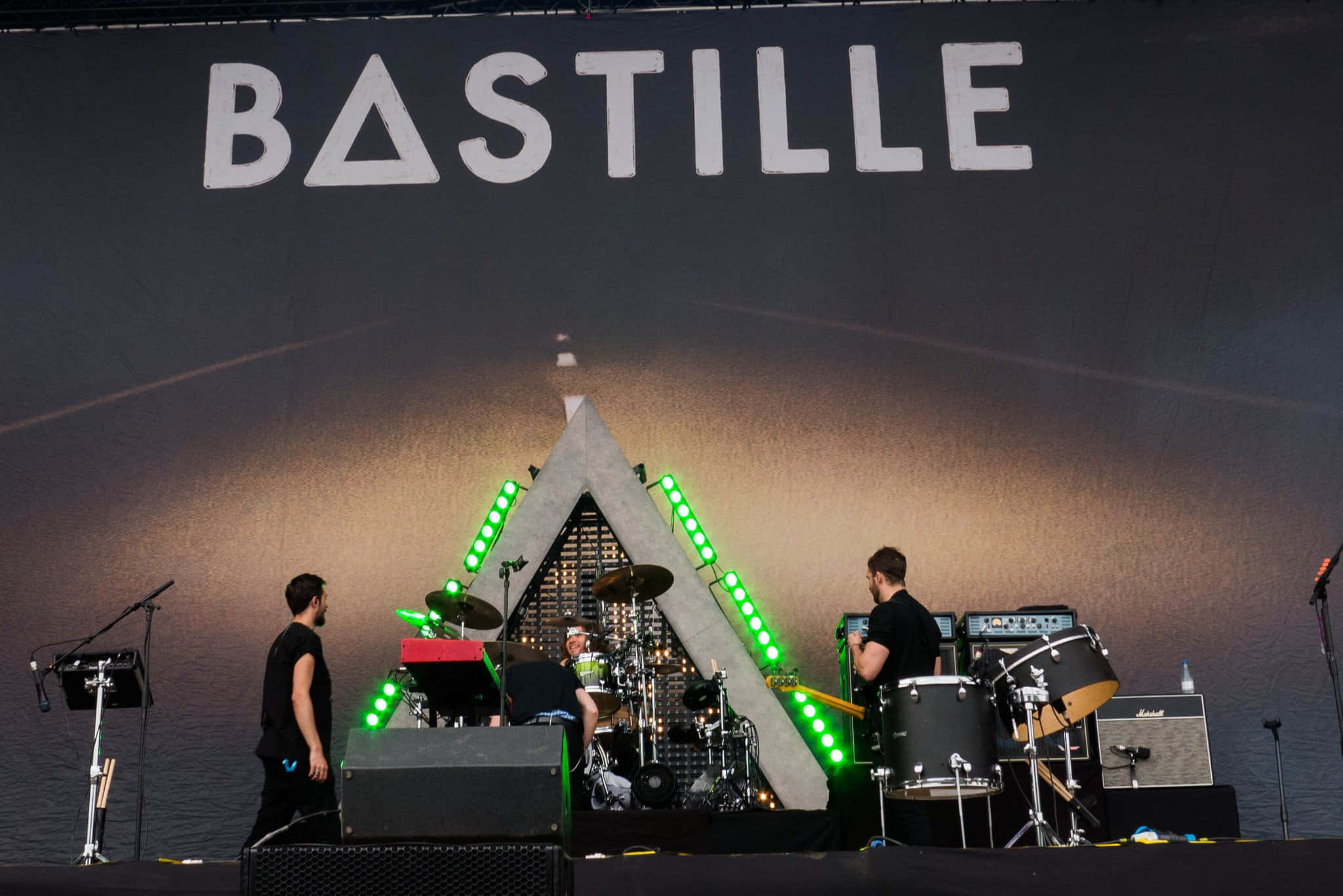 感受Bastille的節奏 壁紙 — Bastille 樂隊在舞台上現場表演，伴隨著發光的綠色燈光和一個大型三角形背景。