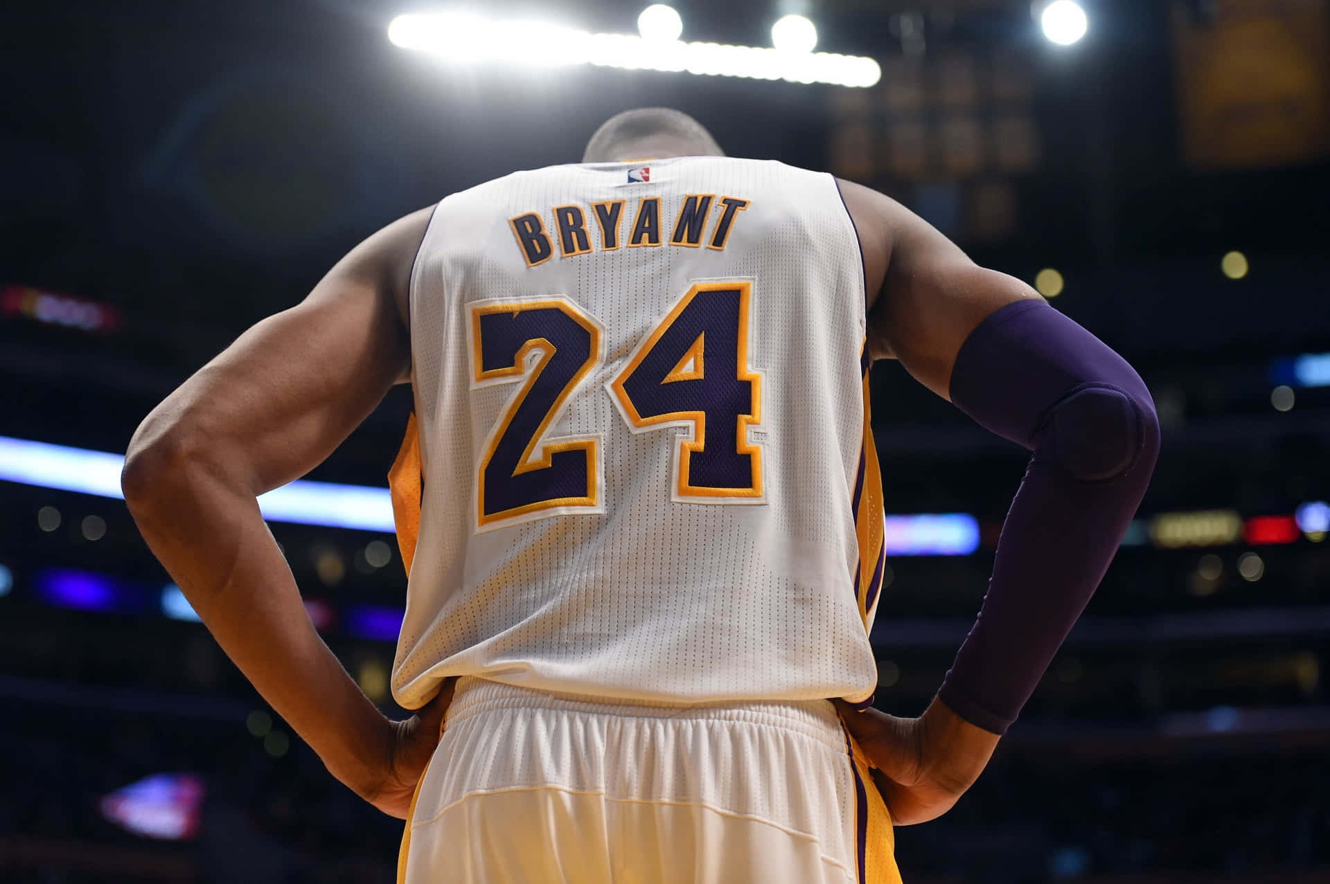感受NBA籃球的熱血激情 壁紙 — 從背後看到身穿白色24號球衣的籃球運動員高比拜仁在場館燈光下手叉腰站立。