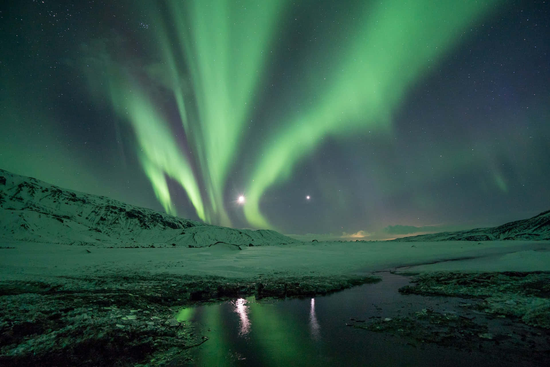 綠色北極光在雪丘和反射著光芒的小溪上方閃爍，星空下。