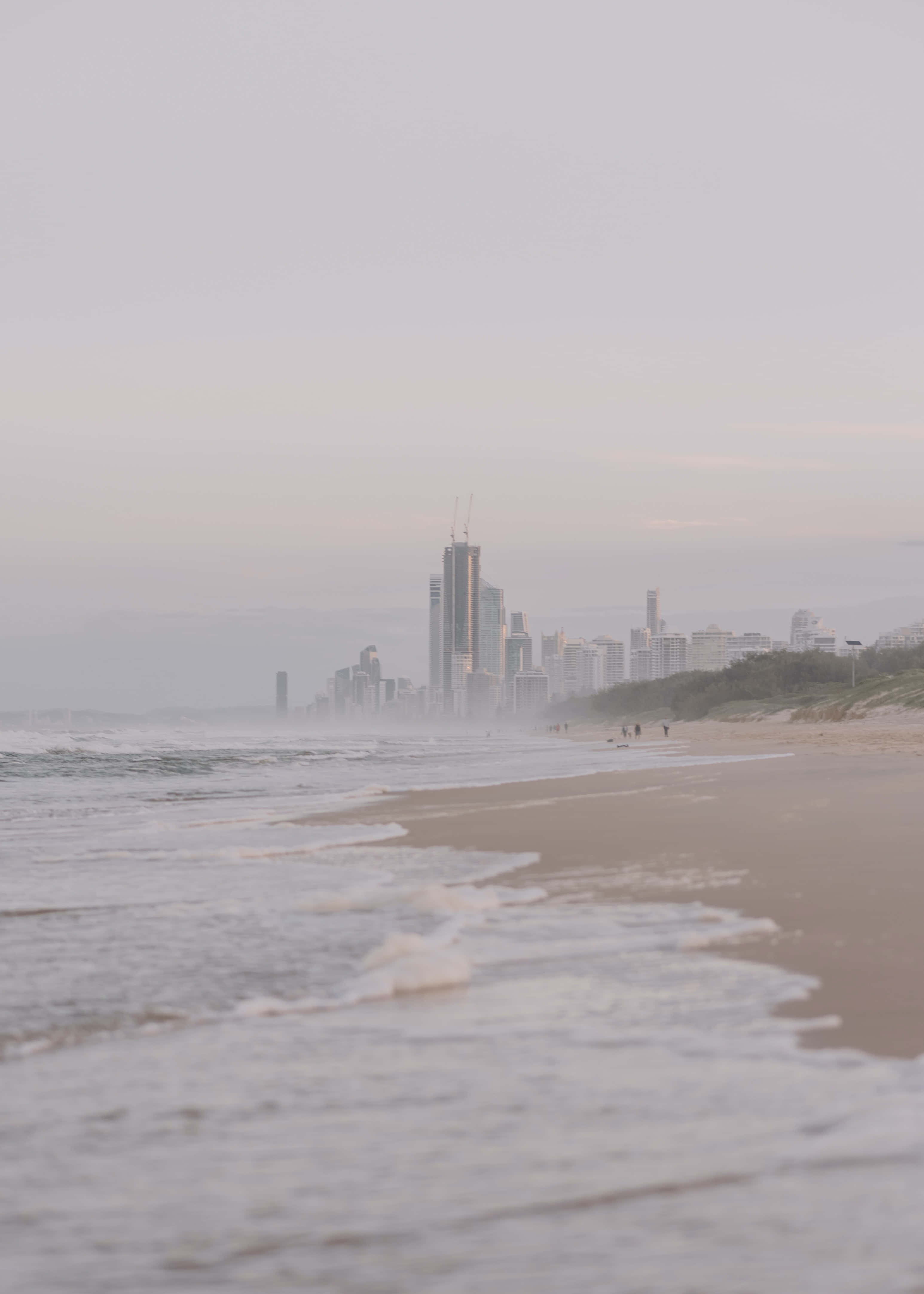 感受海風 - 海灘風情 壁紙 — 霧氣瀰漫的海灘，溫柔的海浪輕拍著岸邊，遠處的城市天際線在朦朧的晨光中漸漸消失。