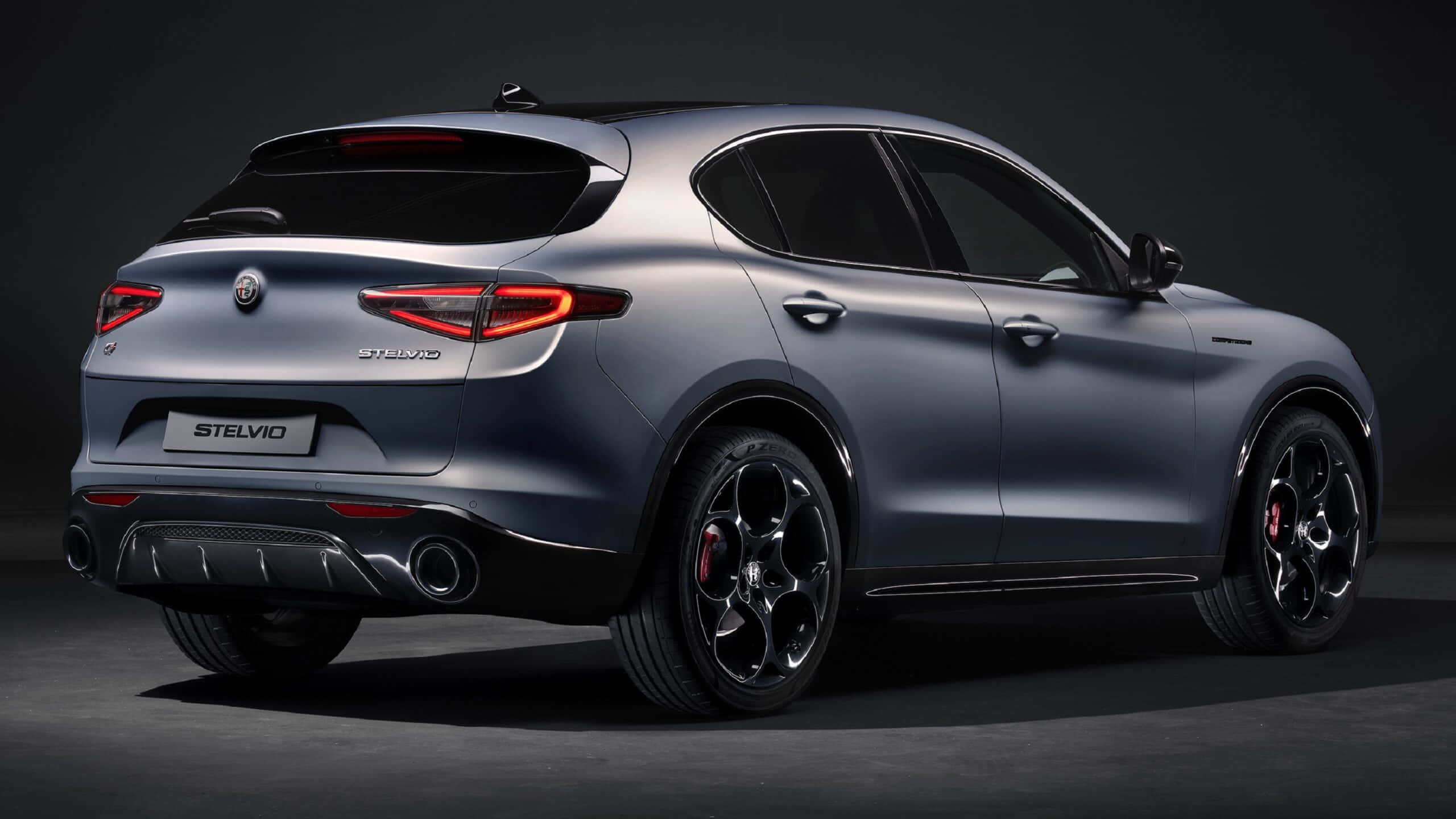 感受義大利熱情，駕馭Alfa Romeo 壁紙 — 一輛時尚的啞光灰色Alfa Romeo Stelvio SUV，從後方四分之三角度展示，帶有發光的紅色尾燈和黑色合金輪圈，背景為深色攝影棚。