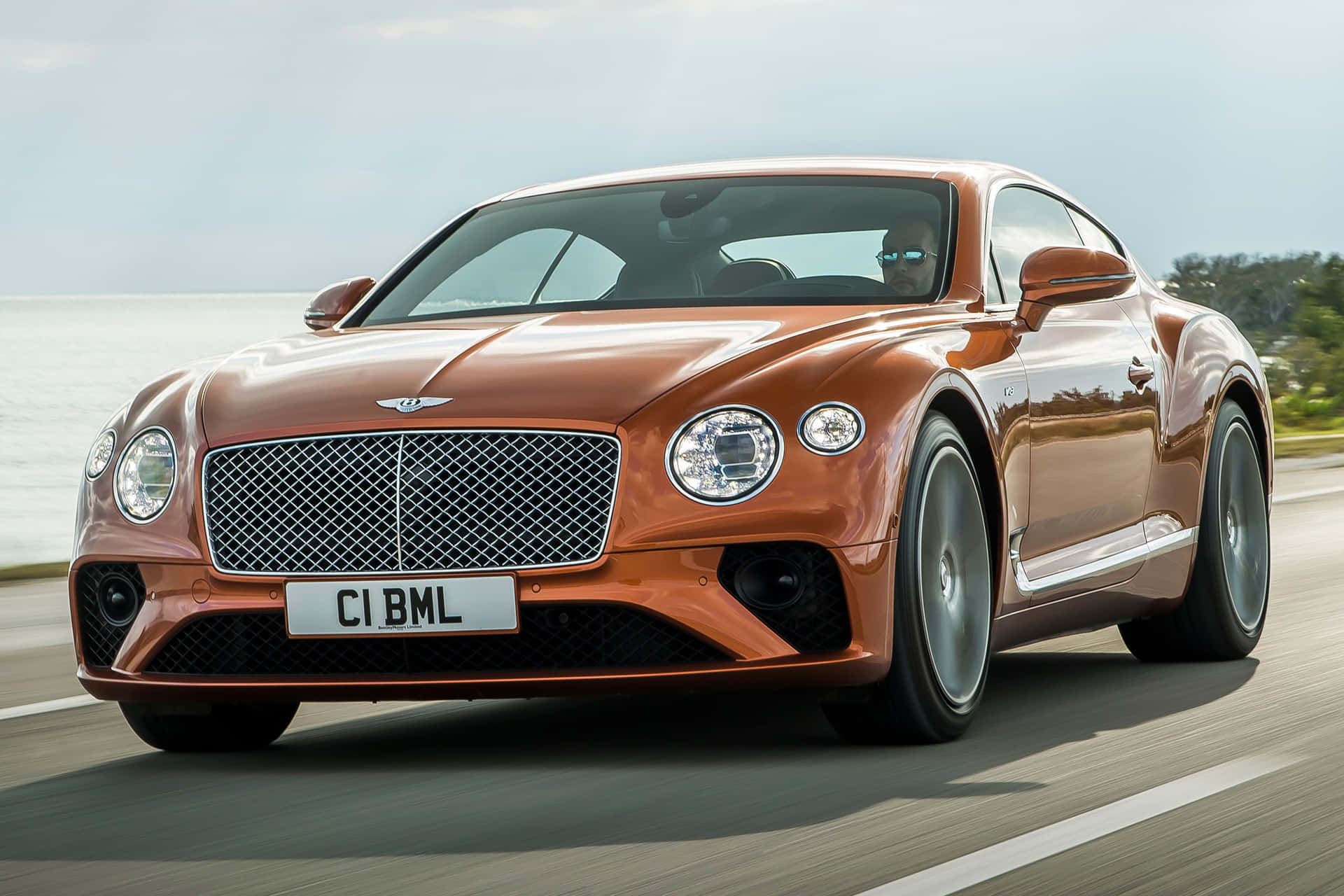 橙色 Bentley Continental GT 在靠近海洋的海岸公路上行駛。