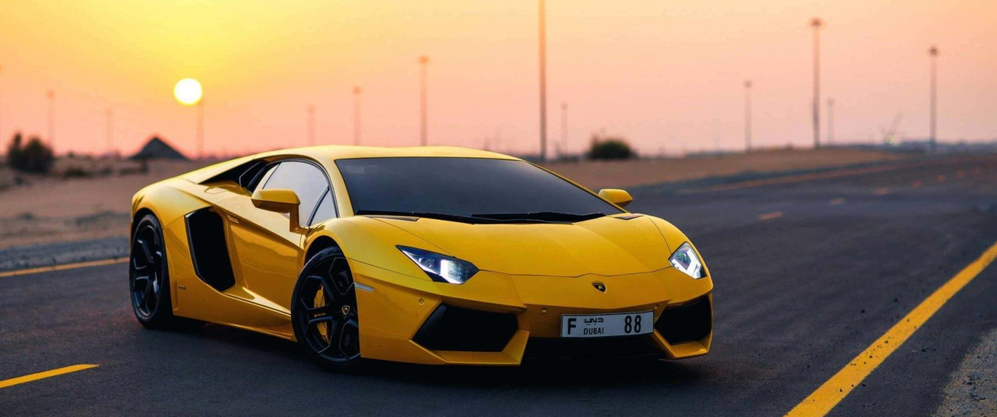 感受這輛絕美蘭博基尼 Aventador SVJ 在開放道路上的澎湃動力 壁紙 — 一輛鮮黃色的林寶堅尼 Aventador 在日落時分沿沙漠公路飛馳，車頭燈閃爍，天空呈現溫暖的橙色。