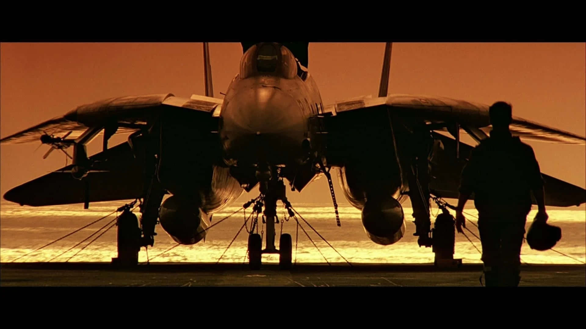感受速度的渴望！ 壁紙 — 一架戰鬥機在航空母艦甲板上以火紅夕陽為背景形成剪影，附近有一名機組人員走過。