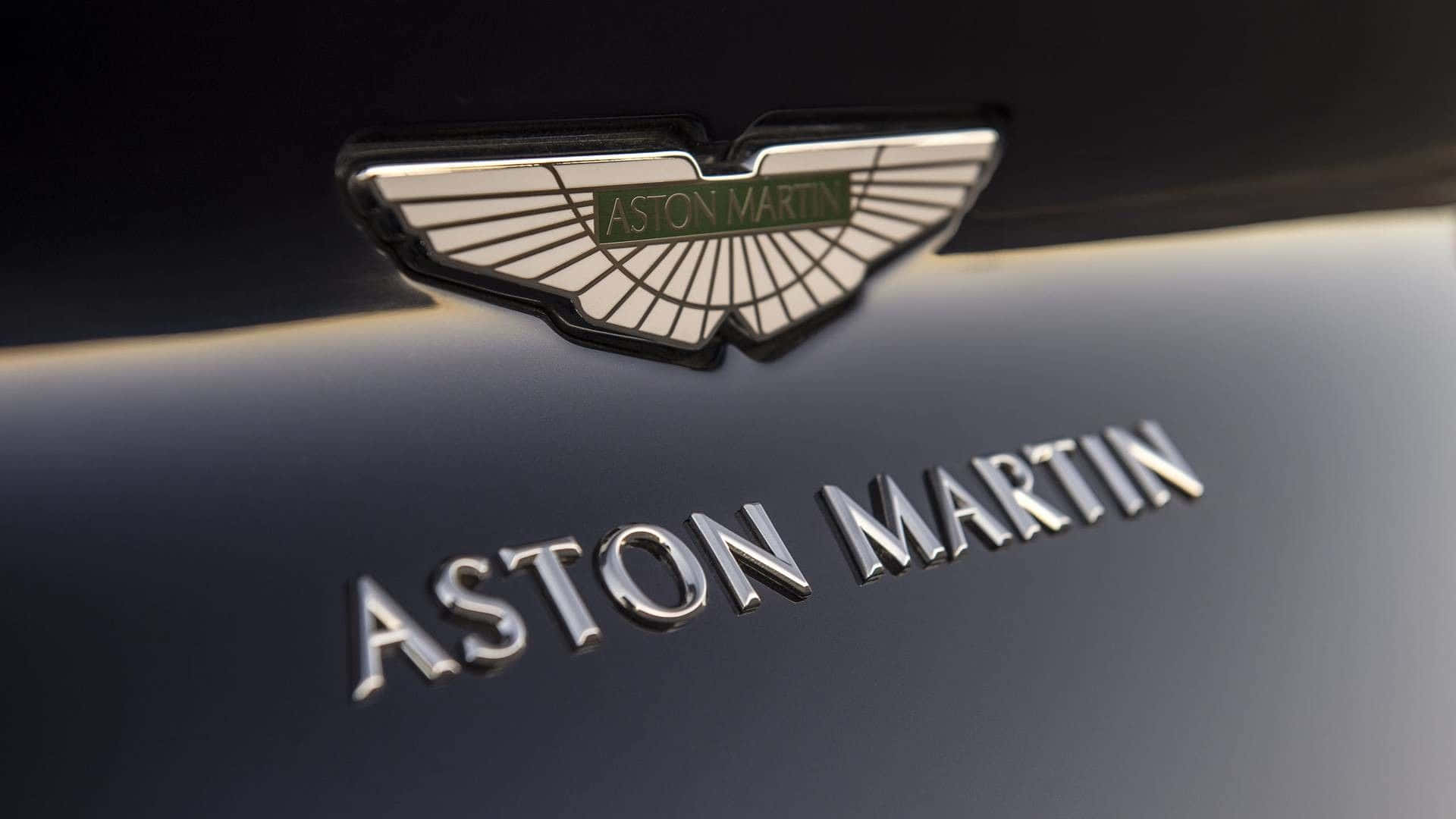 感受阿斯頓·馬丁的力量 壁紙 — 近距離拍攝一個光滑的Aston Martin徽章和字體，位於帶有金屬光澤的深色汽車引擎蓋上。