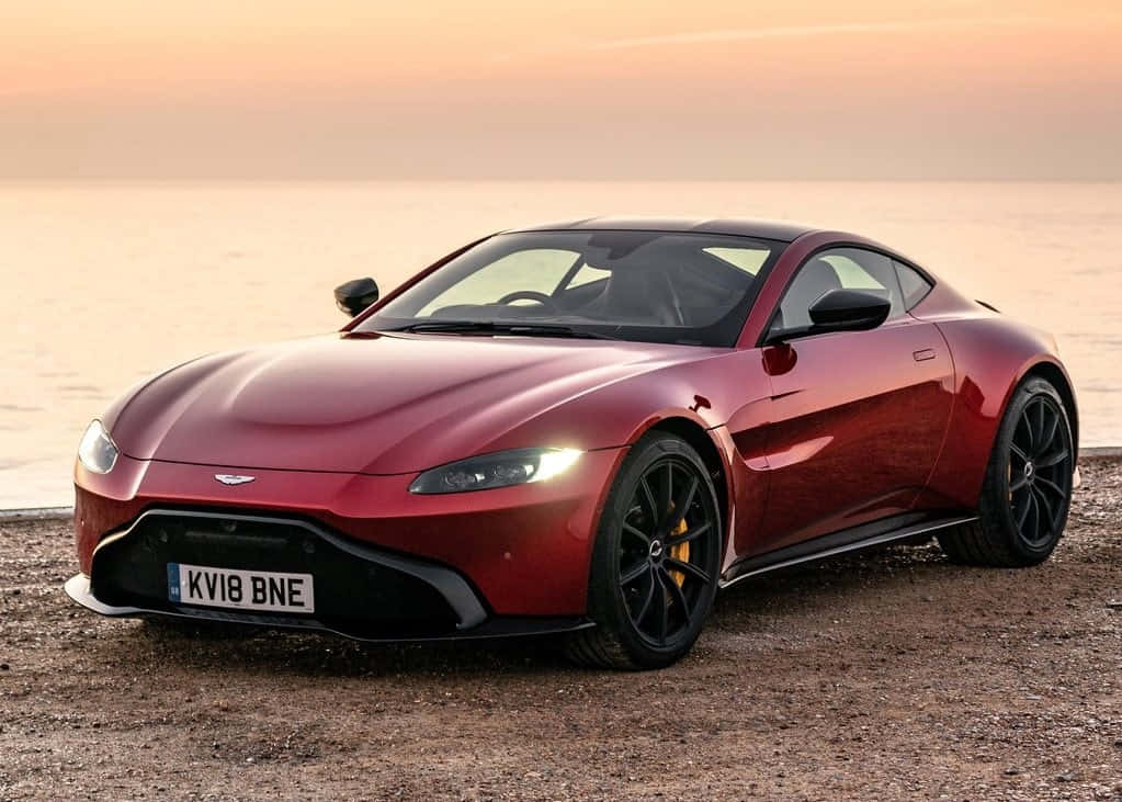 感受駕駛阿斯頓·馬丁的極致快感 壁紙 — 一輛流線型紅色Aston Martin跑車在日落時分，於卵石海灘上閃耀，背後是平靜的海水。