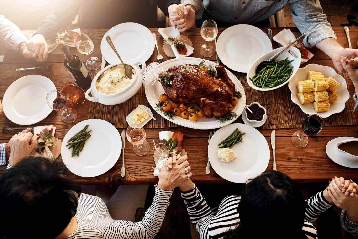 Family gathers around a wooden table for a holiday meal with hands clasped in prayer.