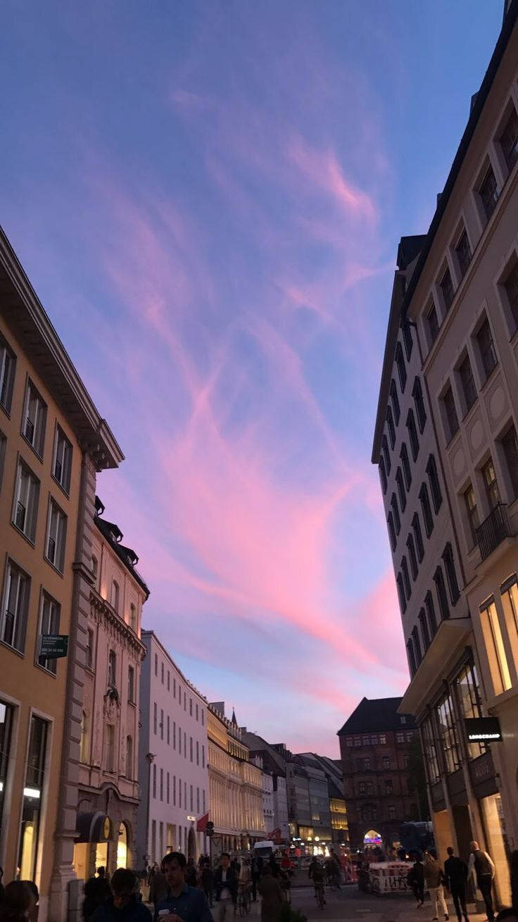 慕尼黑的唯美街景 壁紙 — 黃昏時分的狹窄歐洲街道，粉彩粉紅和藍色雲彩籠罩在歷史建築和下方行人之上。