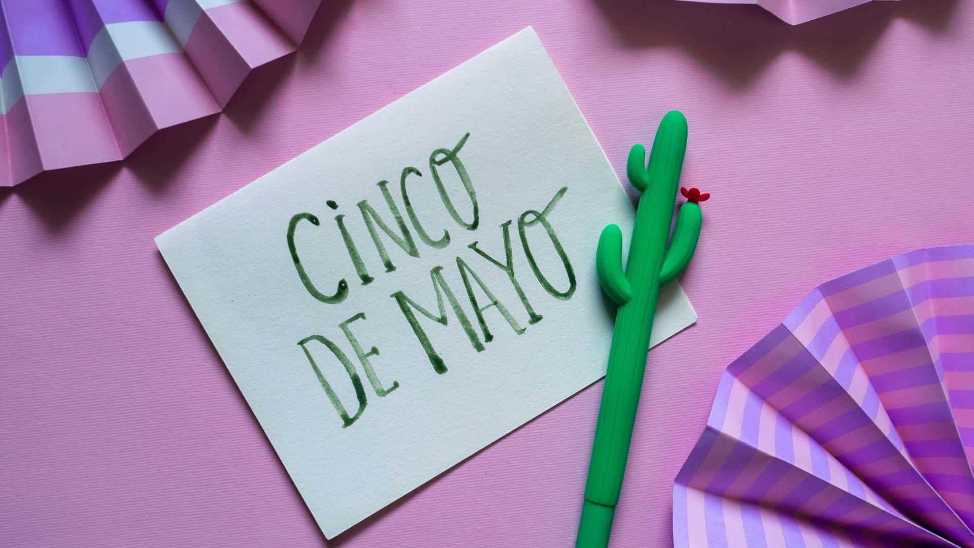 「慶祝五五節！」 壁紙 — 一張手寫的 Cinco de Mayo 便條放在粉紅色表面上，旁邊有綠色仙人掌筆和紫色條紋紙扇。