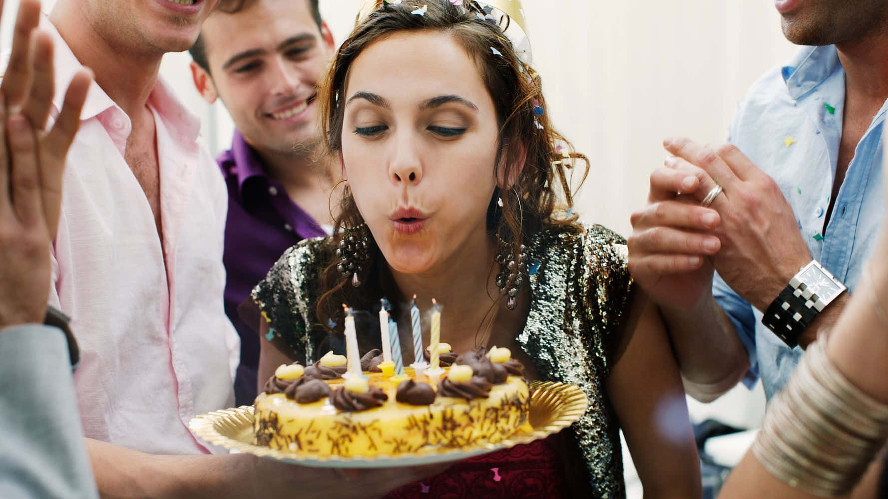慶祝又一年的精彩人生！ 壁紙 — 一位年輕女性在生日派對上吹熄朱古力蛋糕上的蠟燭，朋友們在她周圍歡呼。
