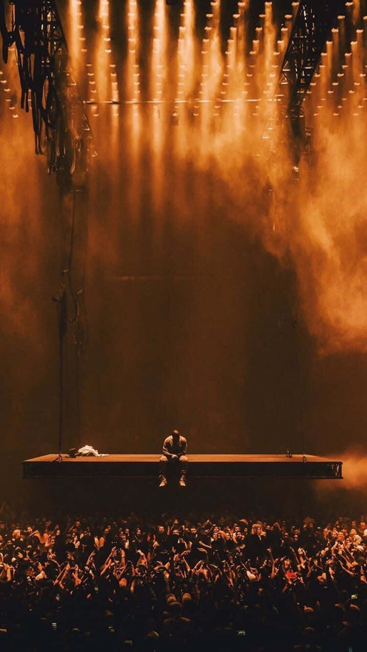慶祝坎耶·威斯特的傑作《The Life Of Pablo》 壁紙 — 一位孤獨的表演者坐在舞台中央，沐浴在金色聚光燈下，俯瞰一片歡呼的粉絲海洋。
