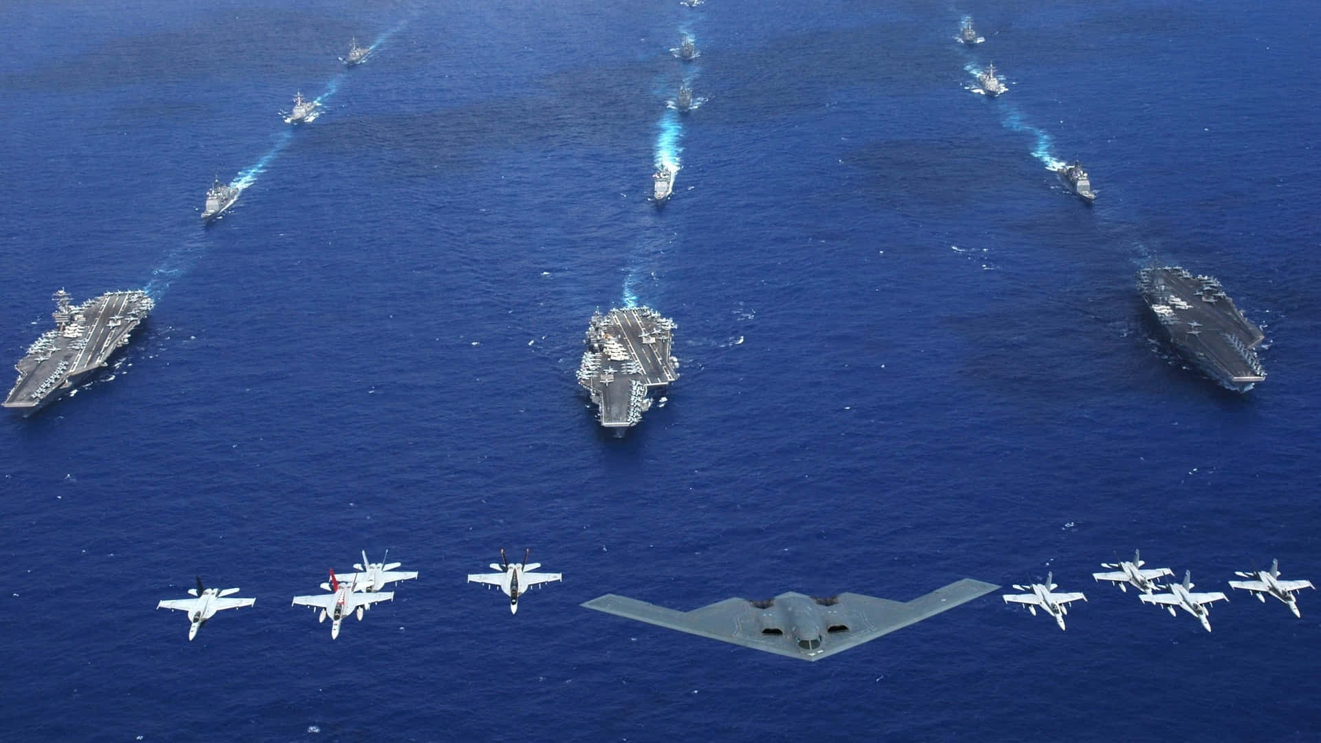 慶祝美國海軍的力量與保護力 壁紙 — 三艘航空母艦及戰鬥機在深藍色海洋上空編隊飛行的鳥瞰圖。