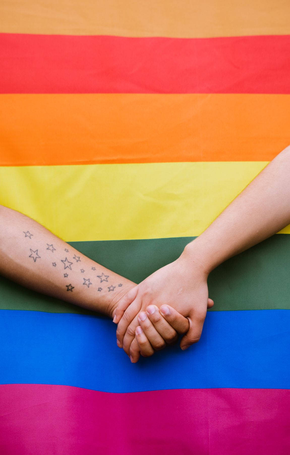 慶祝隱晦LGBT意識 壁紙 — 兩隻手緊握在一起，置於鮮豔的彩虹旗之上，其中一隻手臂上有星星紋身。