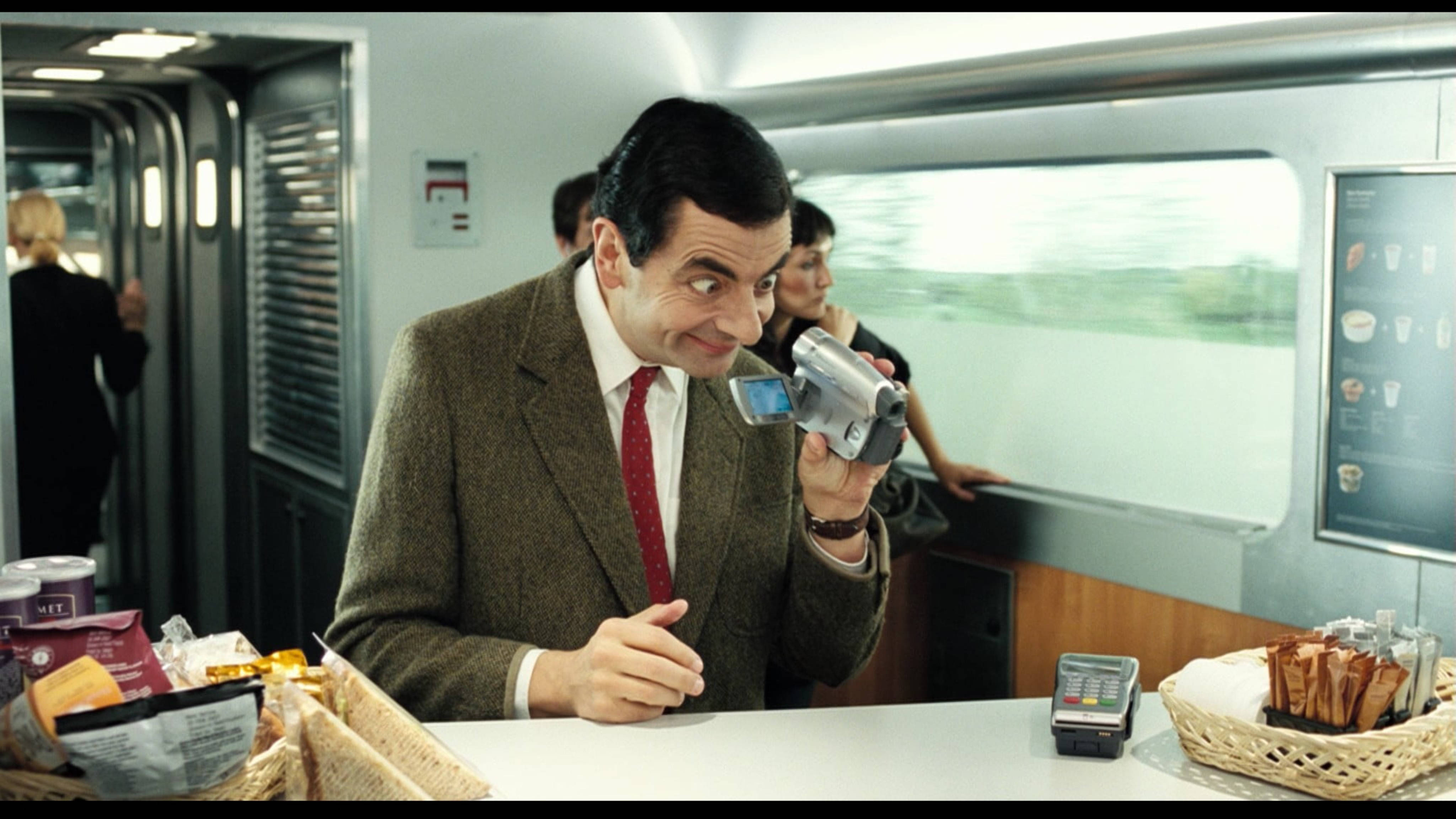 憨豆先生假期偷拍美食 壁紙 — Mr Bean 喺行駛中嘅火車車廂內用輕便攝錄機拍攝自己，露出頑皮嘅笑容。