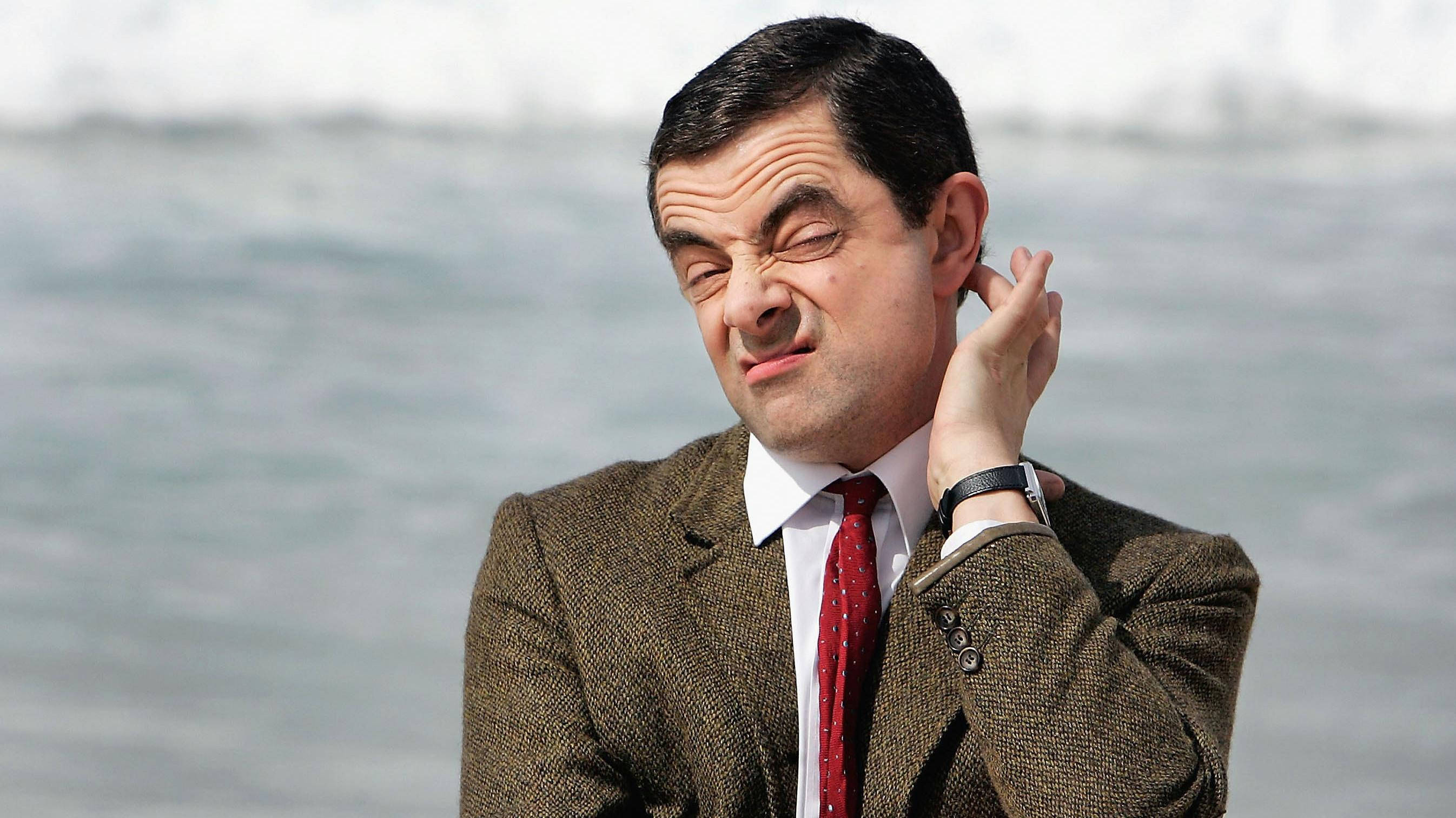 憨豆先生抓頭姿勢 壁紙 — Mr Bean 身穿棕色粗花呢西裝和紅色領帶，在模糊的水背景附近抓脖子，表情痛苦。