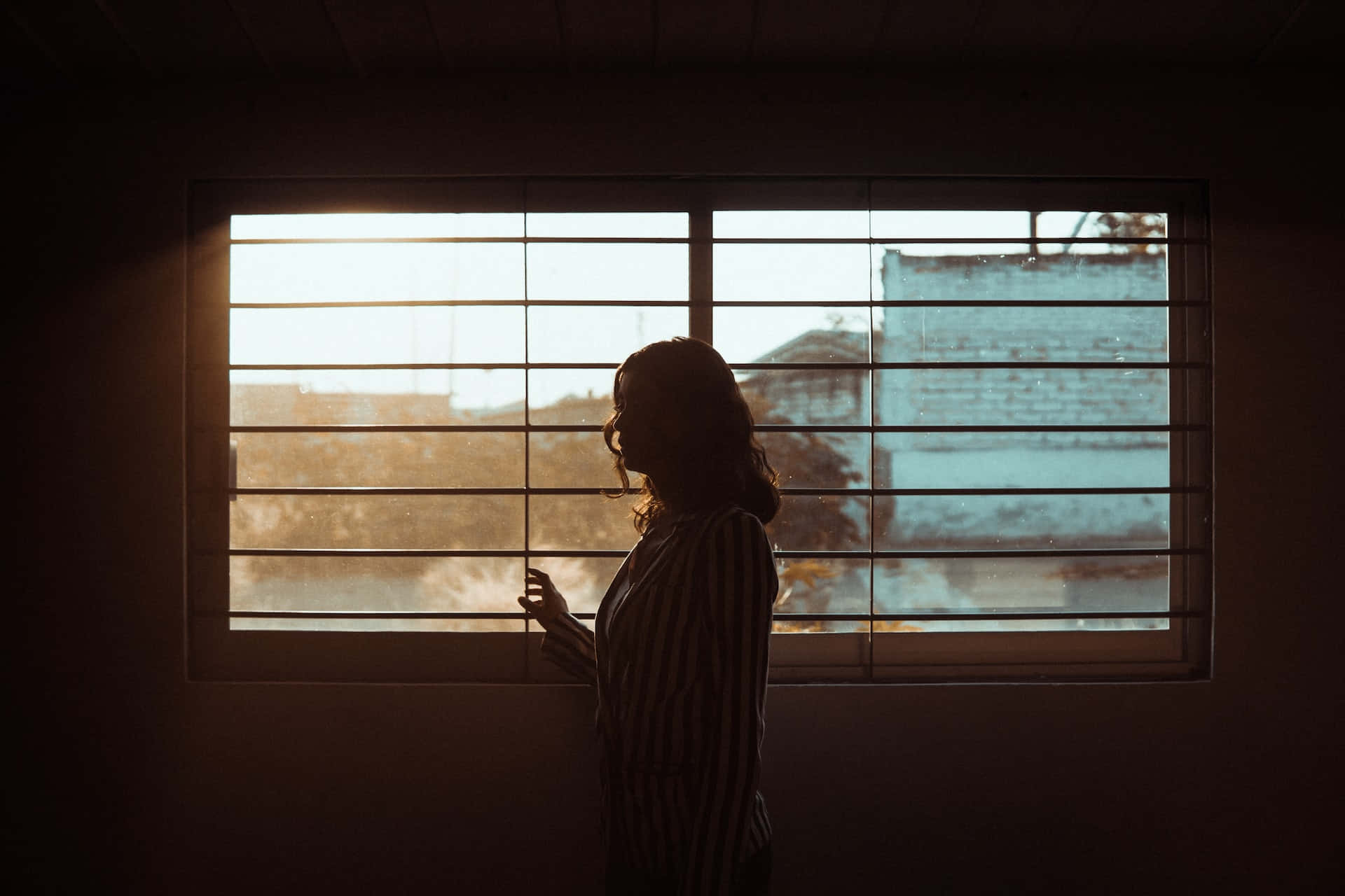 女人剪影在黃金時刻透過百葉窗凝視窗外。
