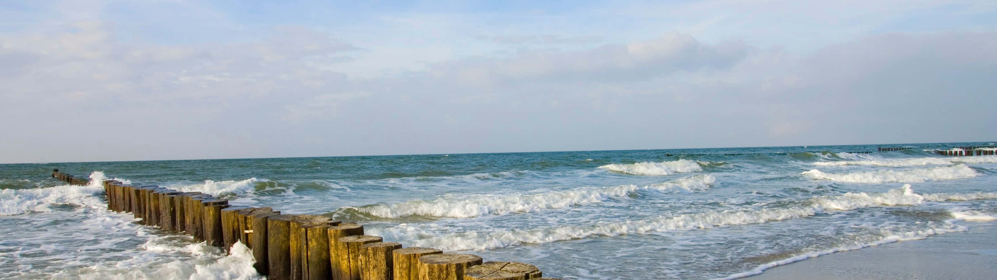 懸崖邊的湛藍海景 壁紙 — 海浪拍打著延伸至廣闊藍海的風化木製防波堤，天空柔和多雲。