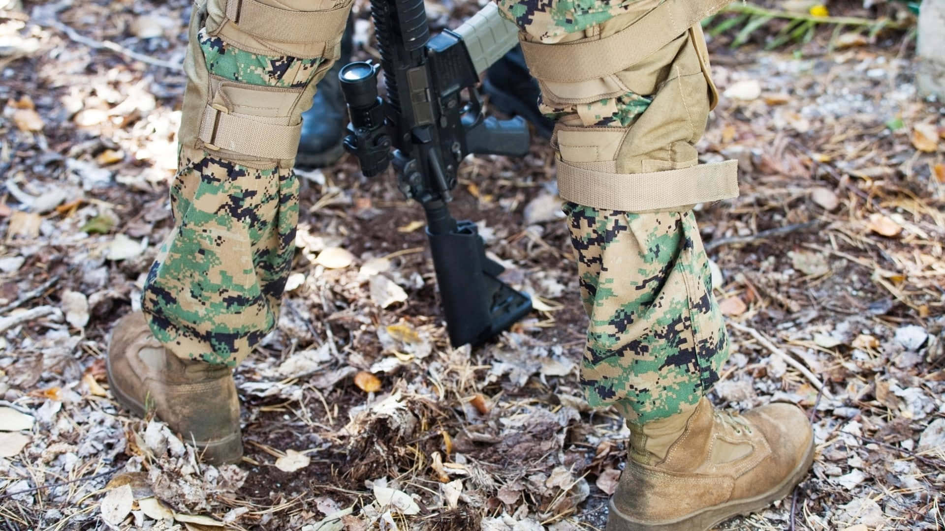 成為軍隊所需的英雄 壁紙 — 士兵穿著迷彩裝備，在森林地面的落葉中手持步槍的腿部特寫。
