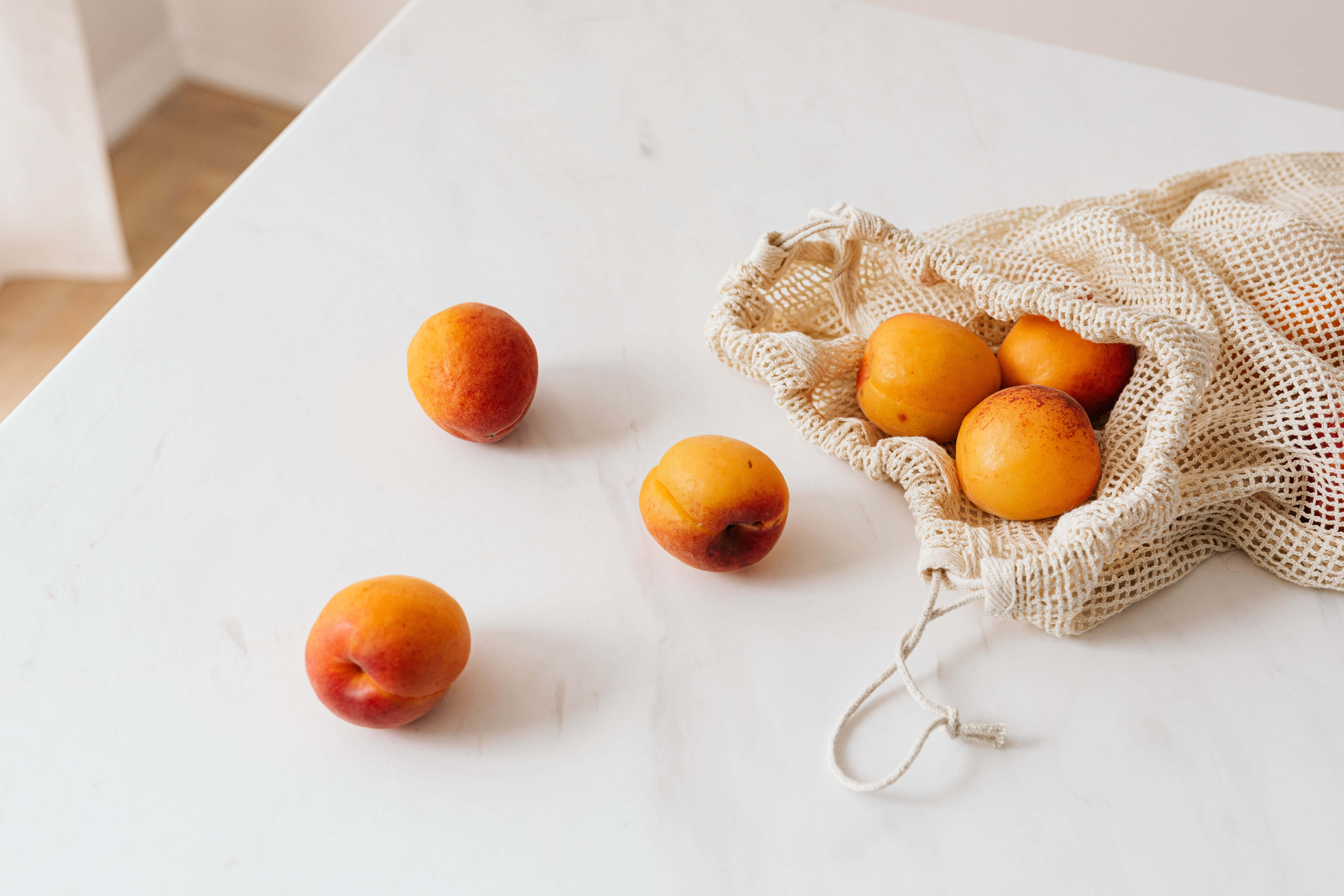 Summer Fruits 桌布 壁紙 — 成熟的杏子從米色網袋中溢出，落在柔和的自然光下潔淨的白色大理石檯面上。