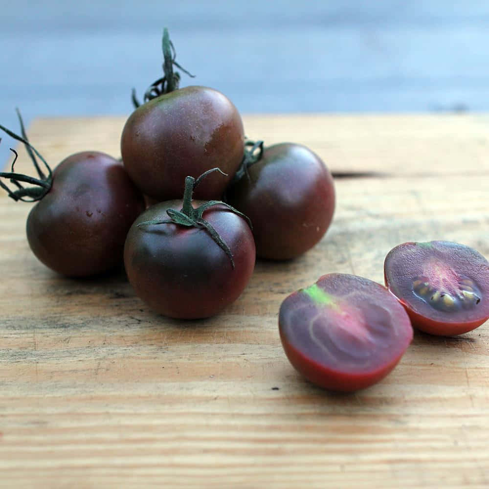 Black Cherry 桌布 壁紙 — 成熟的深紫色番茄帶綠色莖部放在一塊風化的木板上，其中一個切開一半，露出粉紅色的果肉。