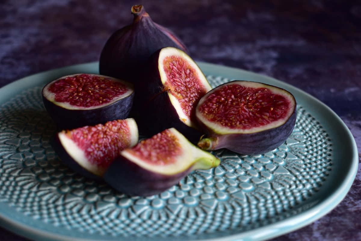 Purple Figs 桌布 壁紙 — 成熟的紫色無花果切開，露出多汁的紅色果肉，放在有紋理的青綠色盤子上，背景為深色。