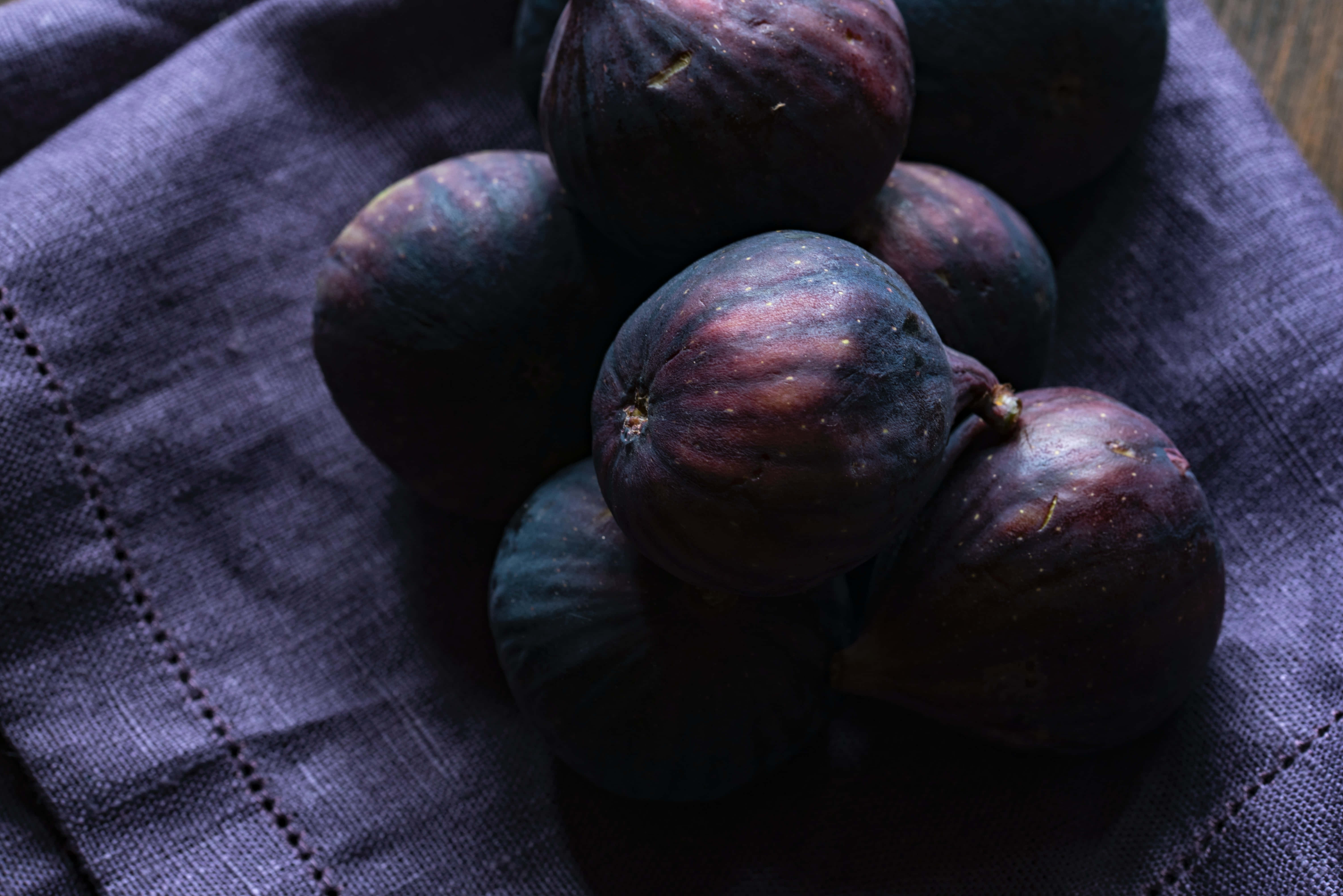 Purple Figs 桌布 壁紙 — 成熟的紫色無花果堆放在有紋理的深紫色布料上，光線柔和。