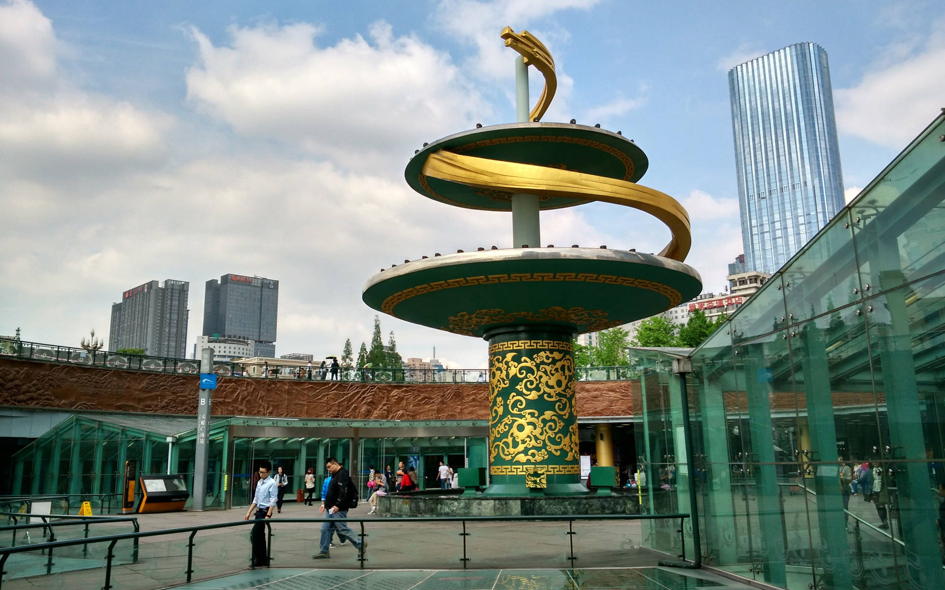 現代城市廣場上的金色螺旋雕塑，有玻璃建築和行走的人們。