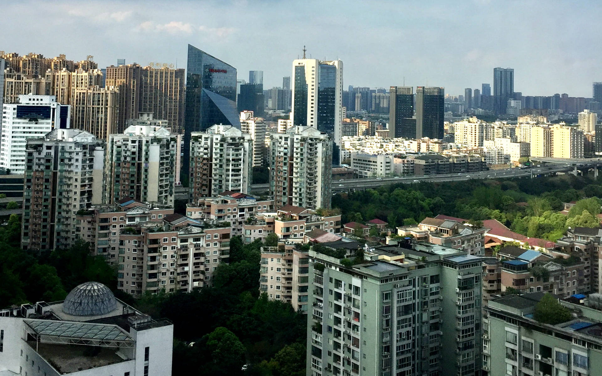 多雲天空下，高樓大廈與綠植密集的城市景觀。