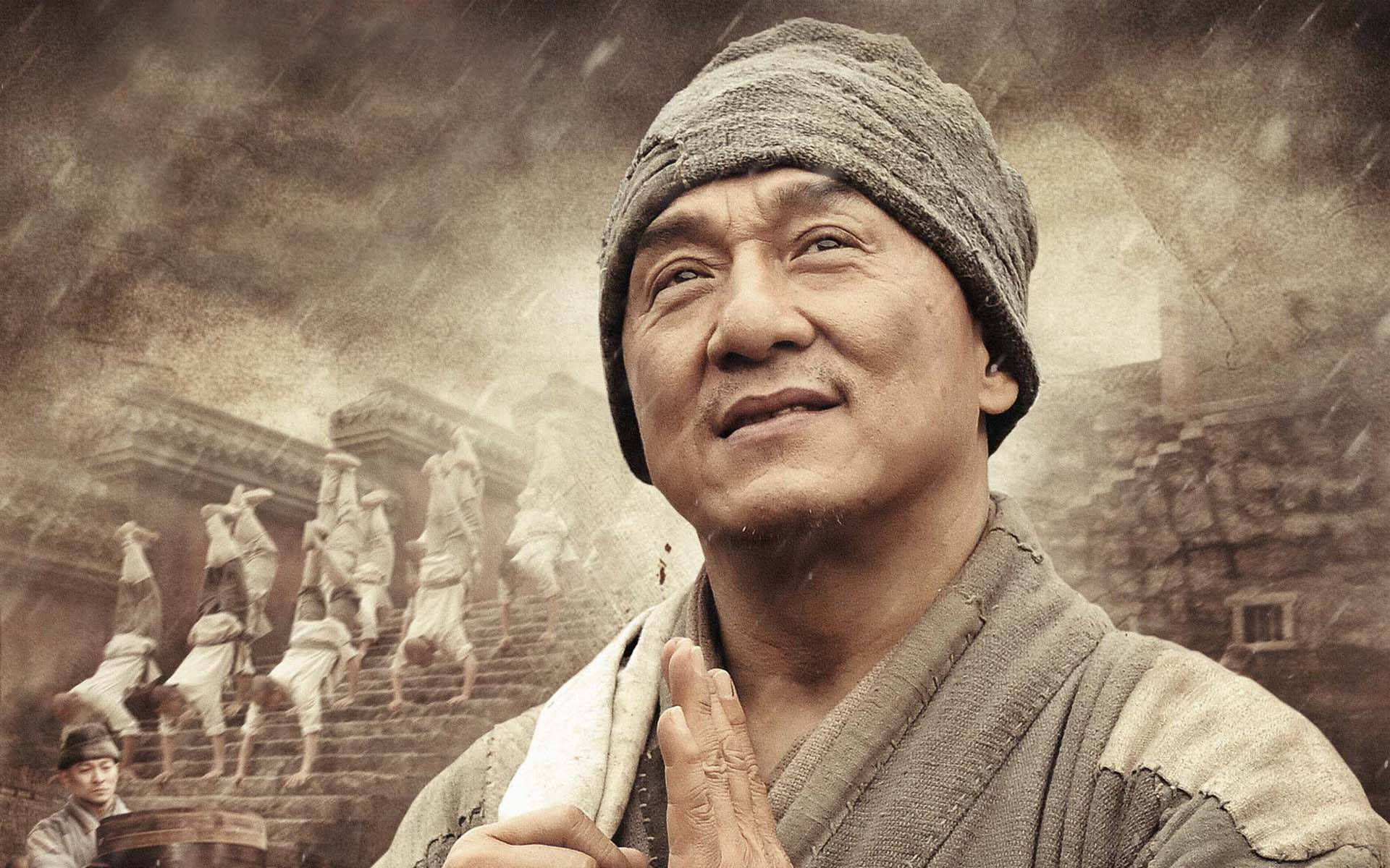 成龍《少林寺》電影海報 壁紙 — 一個戴羊毛帽的男人抬頭仰望，表情平靜，身後有僧侶在霧氣瀰漫的懷舊色調場景中登上石階。