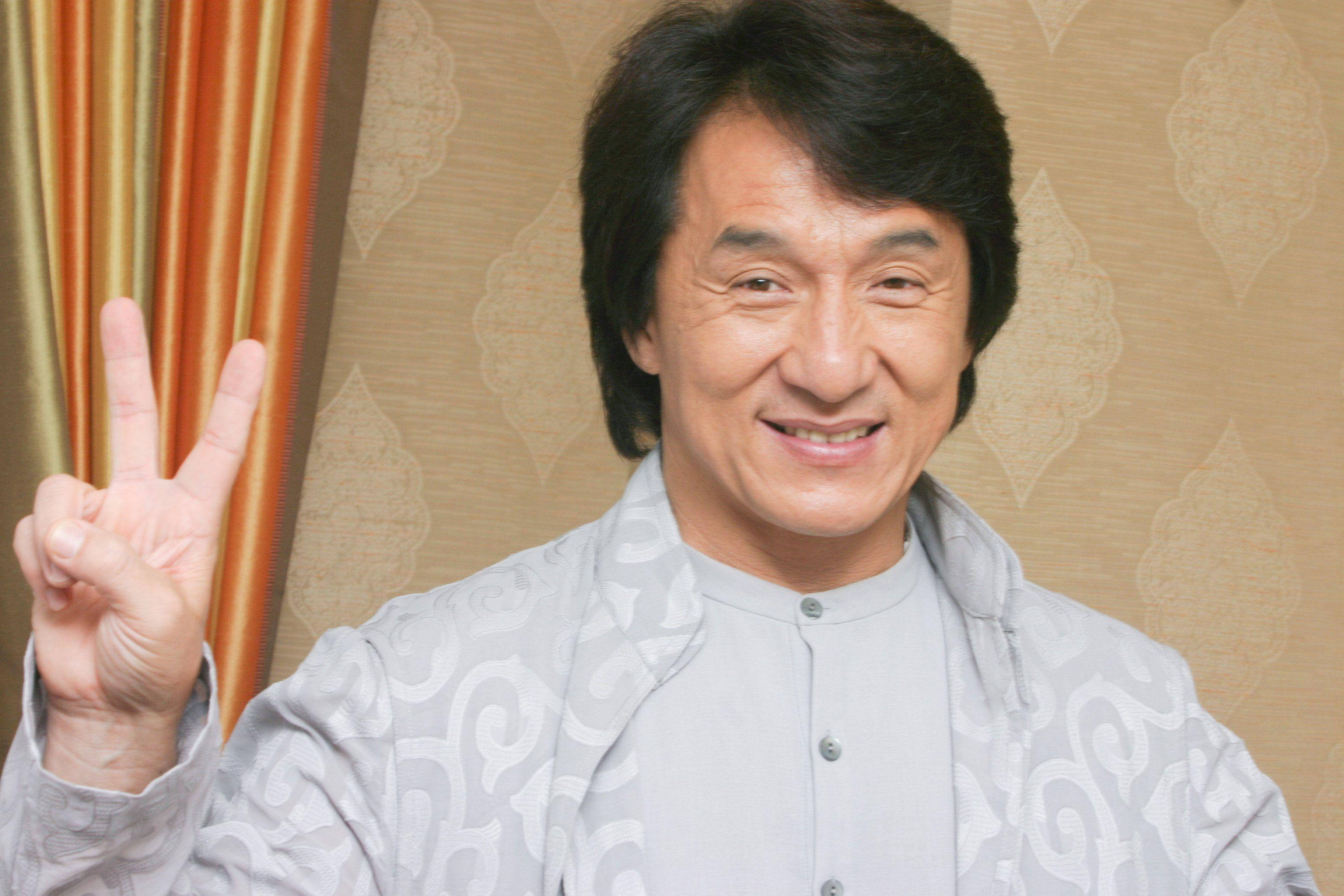 成龍 招手致意 壁紙 — 一位開朗的黑髮男子在圖案牆紙和條紋窗簾前微笑著做出和平手勢。