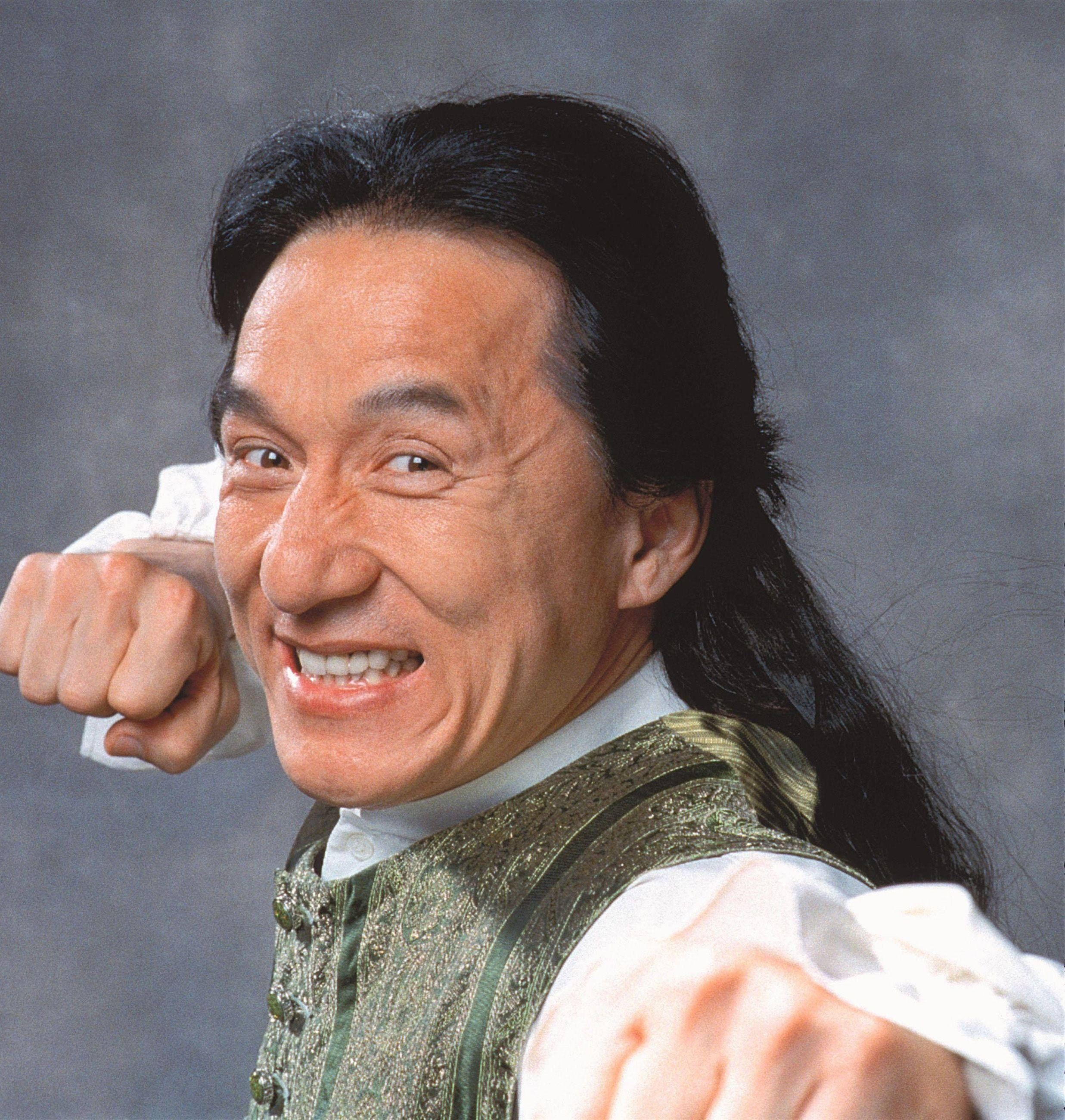 成龍半馬尾造型 壁紙 — 一名長黑髮、穿綠色背心的男子，在灰色背景前高舉拳頭，咧嘴大笑。